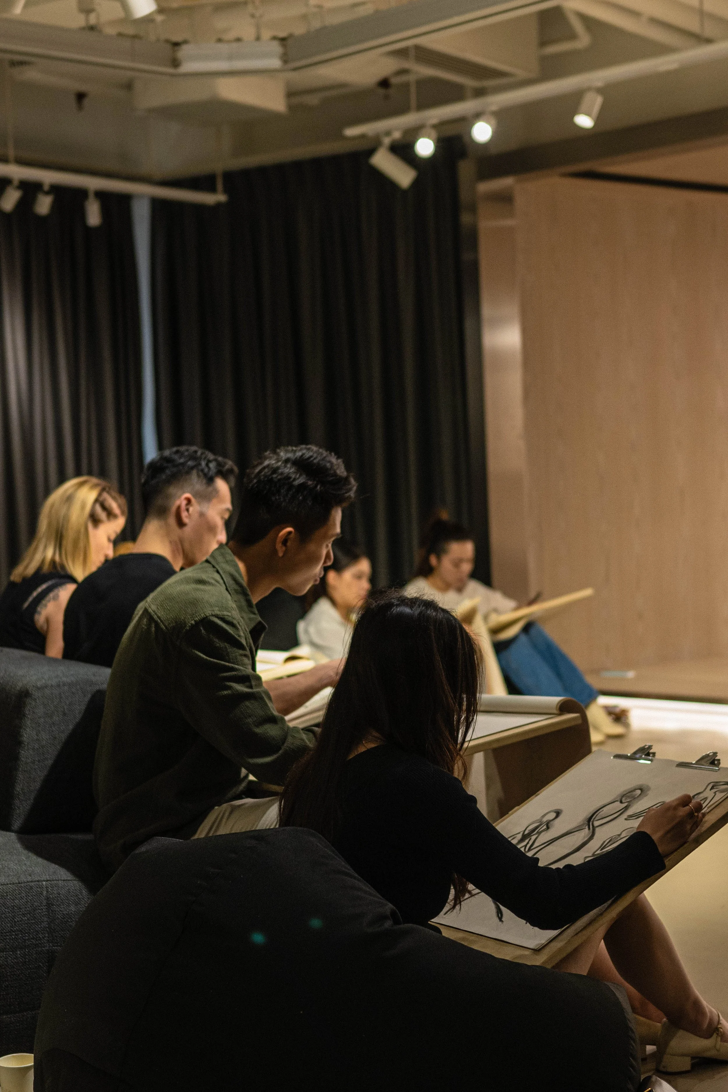 People seated in a row, engaged in a drawing activity, in an indoor setting with a beige wall and black curtains in the background.