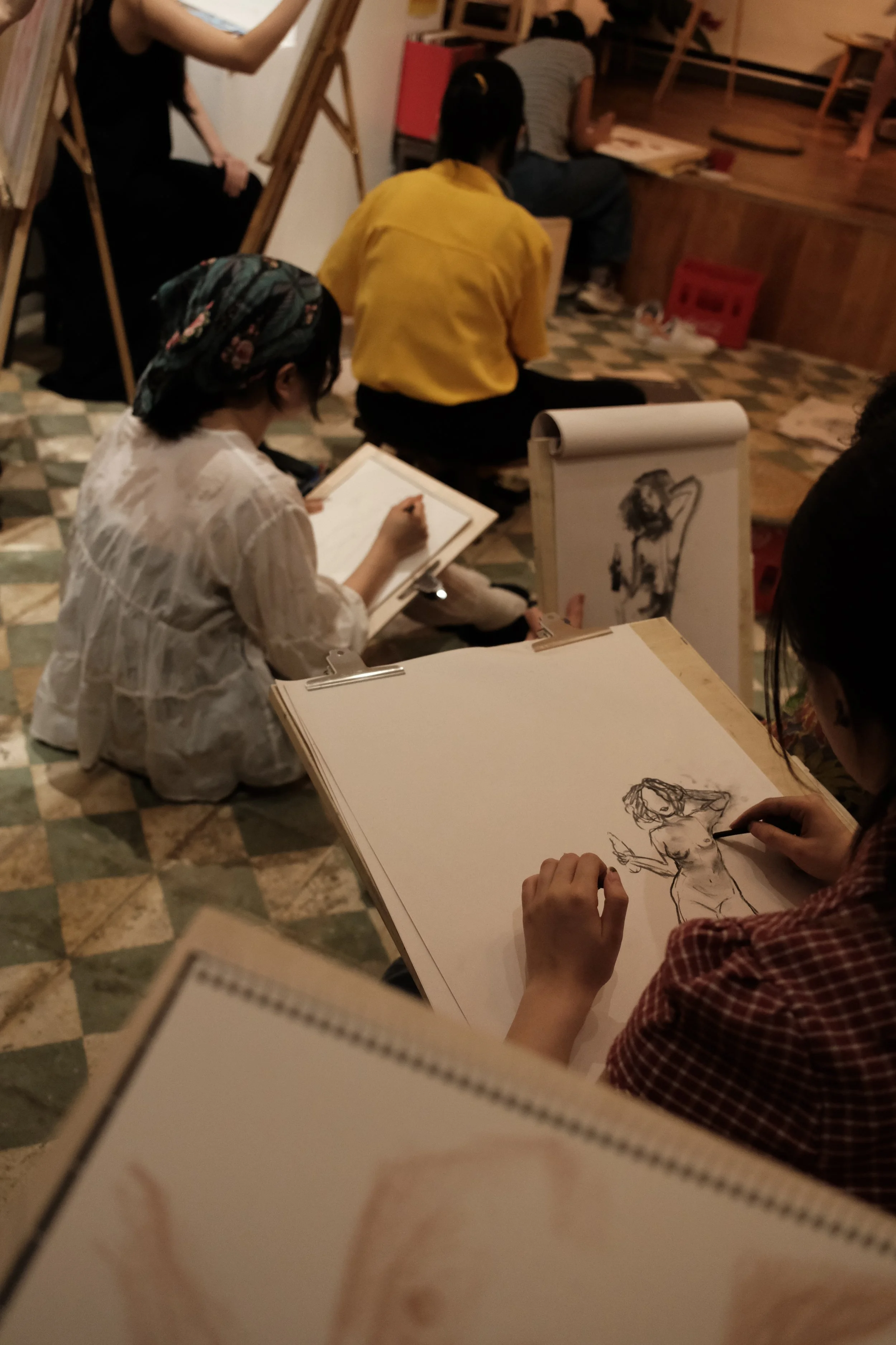 People sitting on the floor and sketching. An artist is drawing a female figure with a strapless top and holding an object. Others are observing or sketching, and some are seated on angled easels or benches.