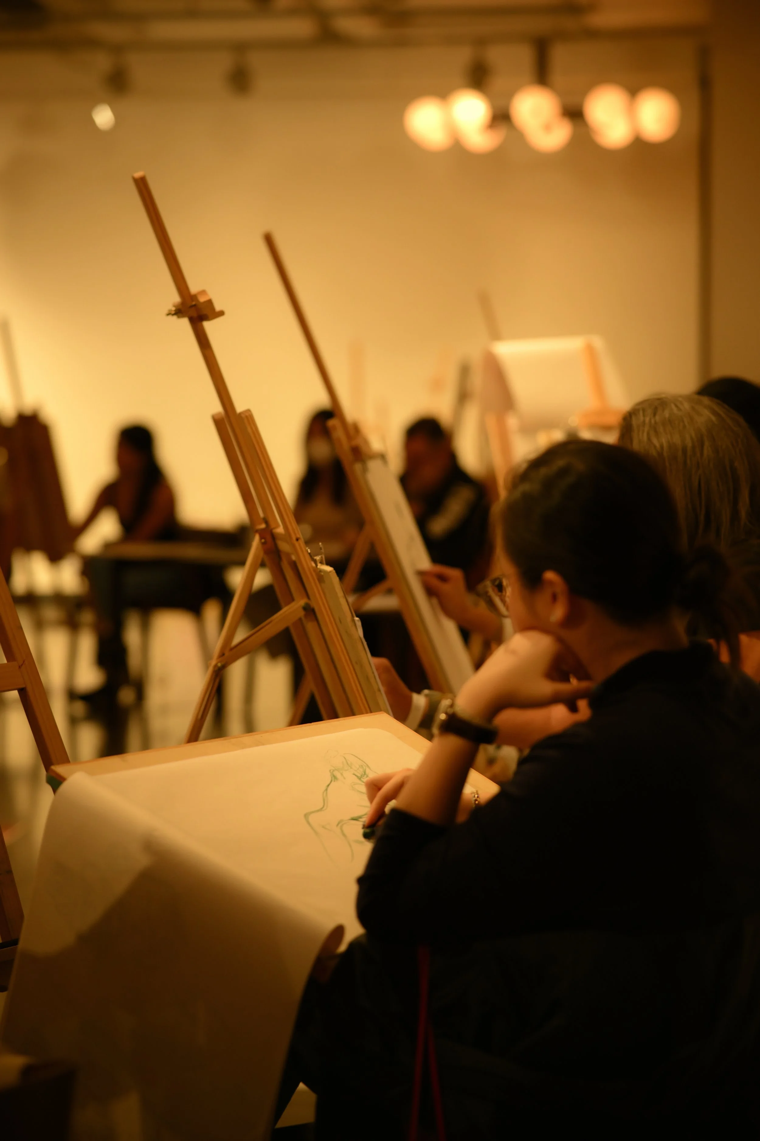 A group of people participating in an art class, drawing on paper with easels in a warmly lit room. The focus is on a woman in the foreground sketching, with several other students and art supplies visible in the background.