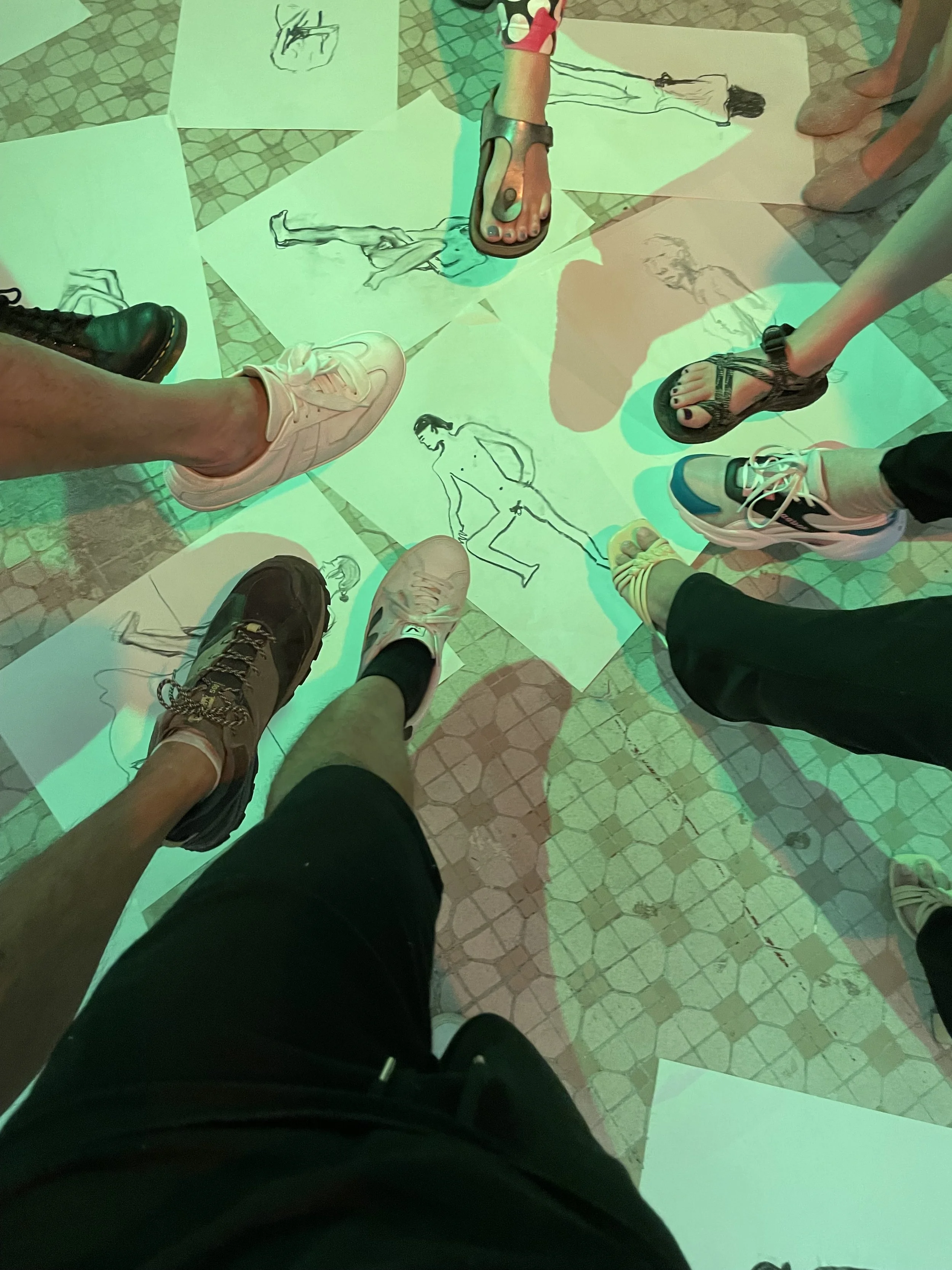 People standing on a flat surface, wearing various types of footwear, with drawings of fashion models on papers scattered on the floor.