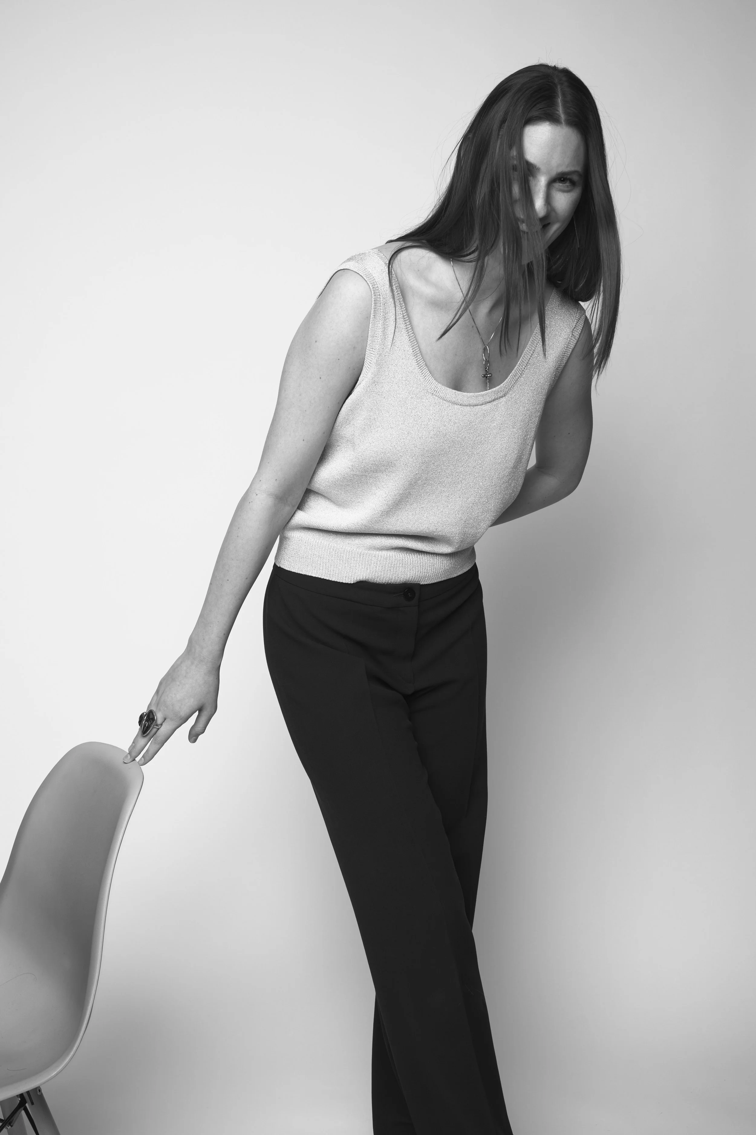 Black and white photo of a woman with long hair smiling and leaning on a chair, wearing a sleeveless top and dark pants, against a plain background.