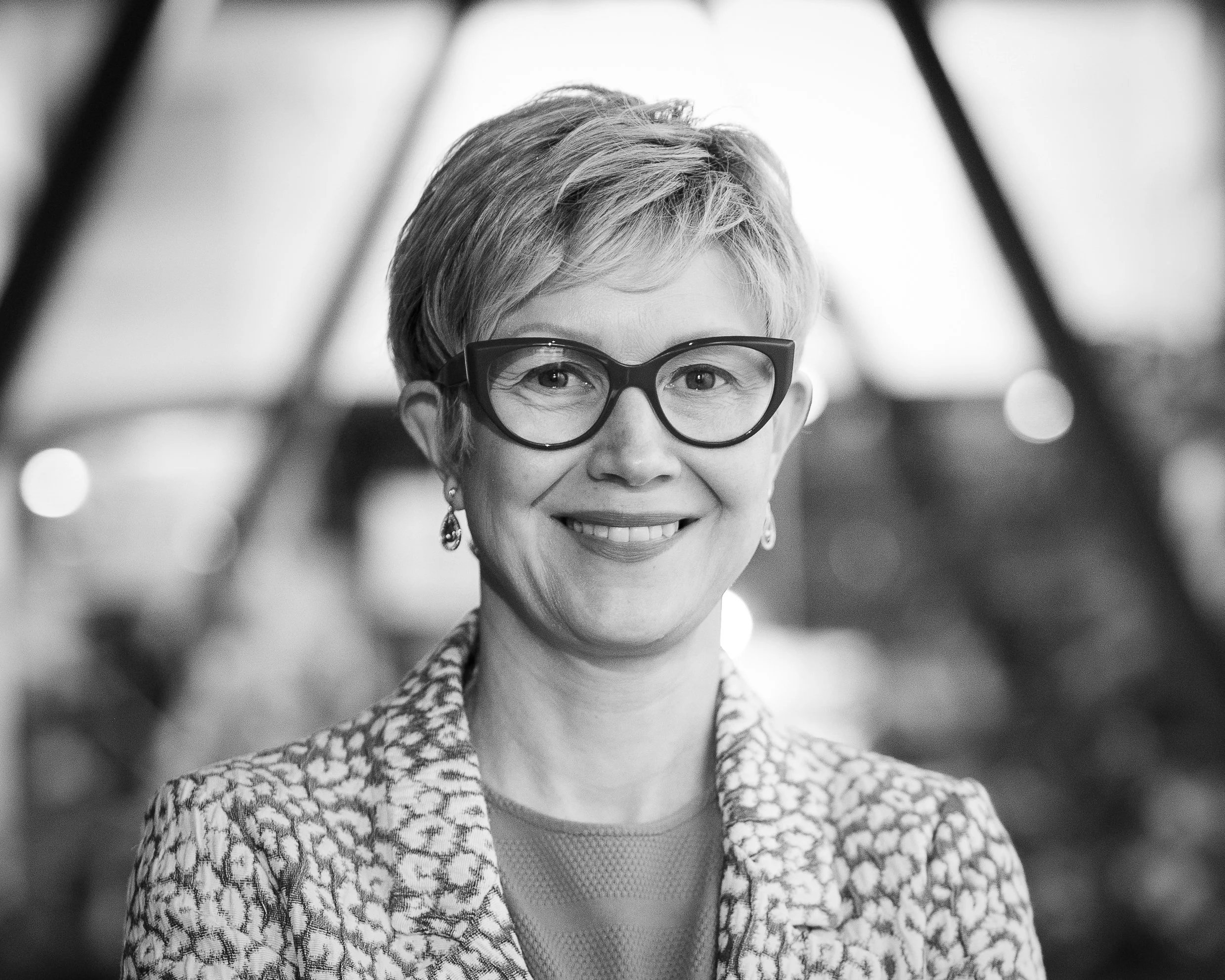 Black and white photo of Miranda Richardson from Willis Bond. She is a woman with short hair, glasses, and earrings, smiling and looking at the camera. She is wearing a patterned blazer over a t-shirt, and the background is blurred with lights.