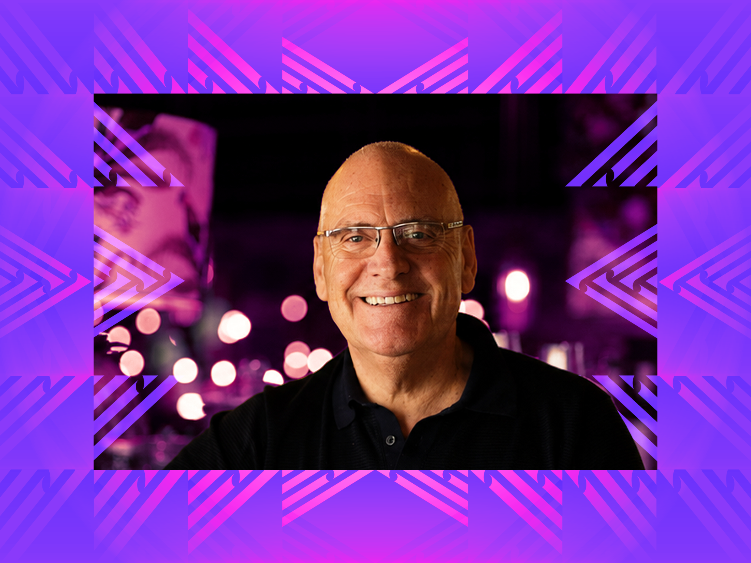 A picture of Mike Egan, owner of Monsoon Poon. He is a smiling man wearing glasses and a black shirt standing in front of a blurred background with purple and pink lights.