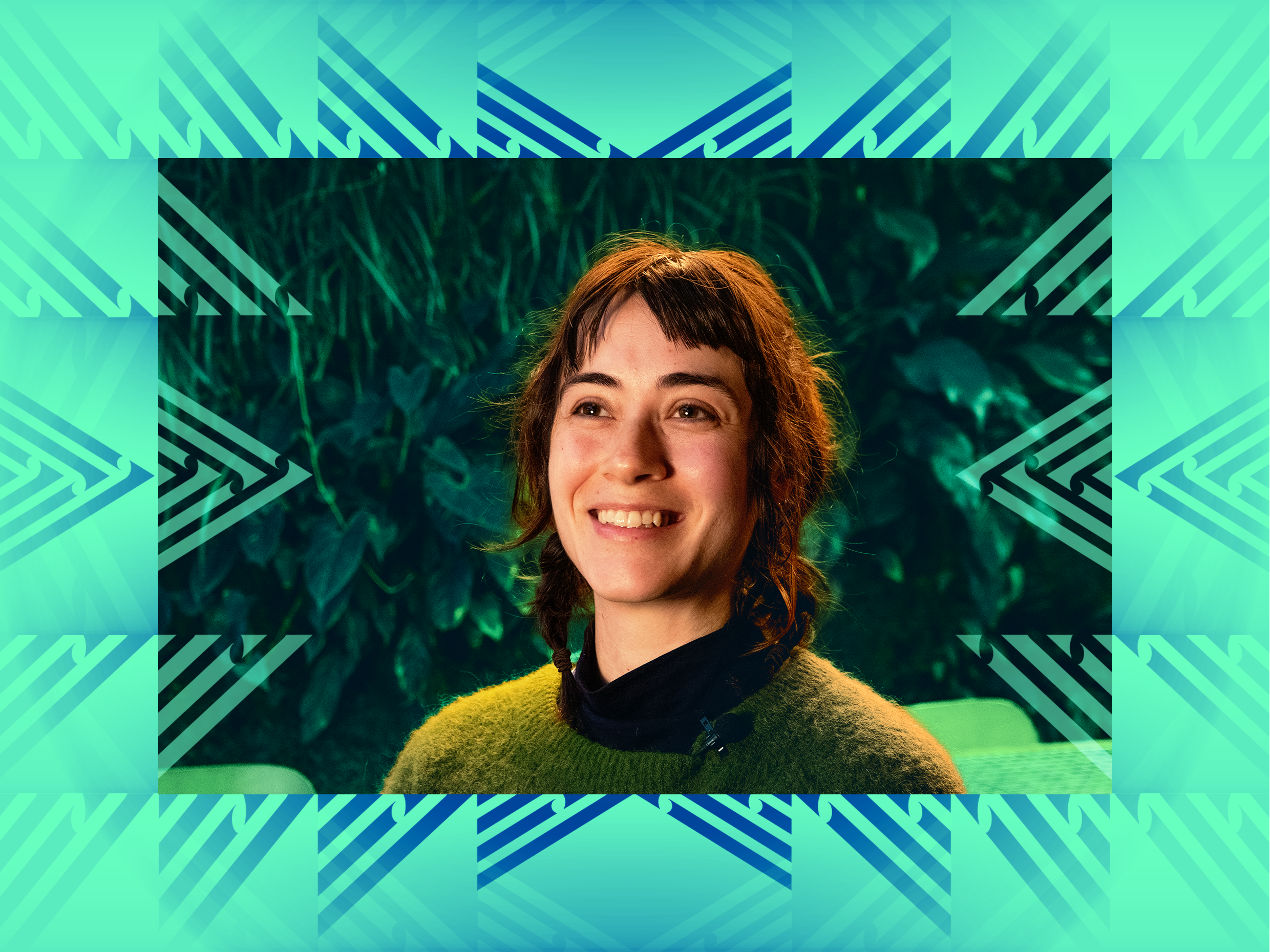 A picture of Flo, another Te Aro Courtenay local. She is a young woman with reddish-brown hair in braids, smiling, wearing a black turtleneck and green fuzzy sweater, sitting in Glou Glou cafe which features a lush greenwall in the background.