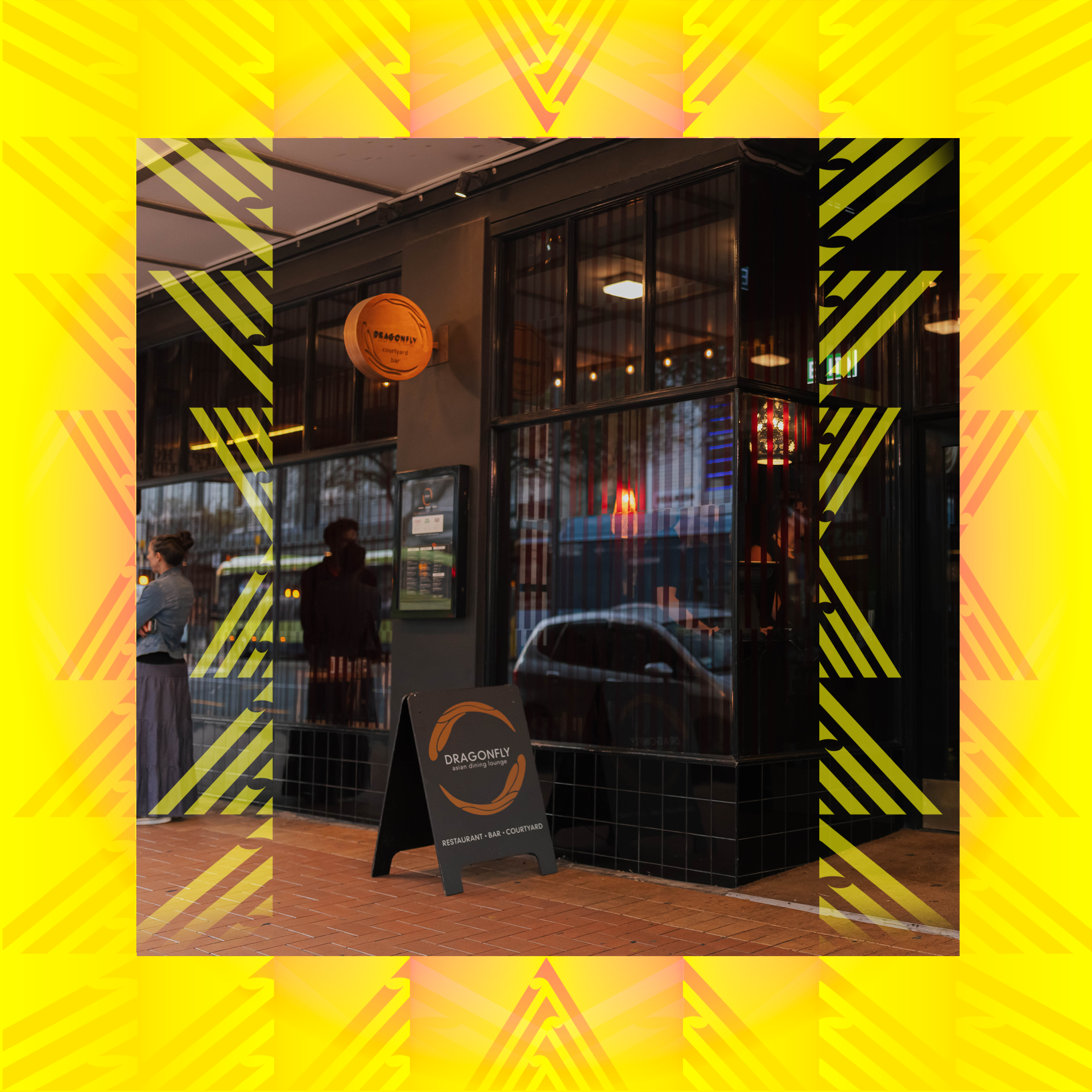 Exterior view of Dragonfly restaurant in Courtenay Place. The building has a black facade and large glass windows, featuring a circular wooden sign for Dragonfly Courtyard Bar and restaurant, and a black A-frame chalkboard sign on a brick sidewalk.
