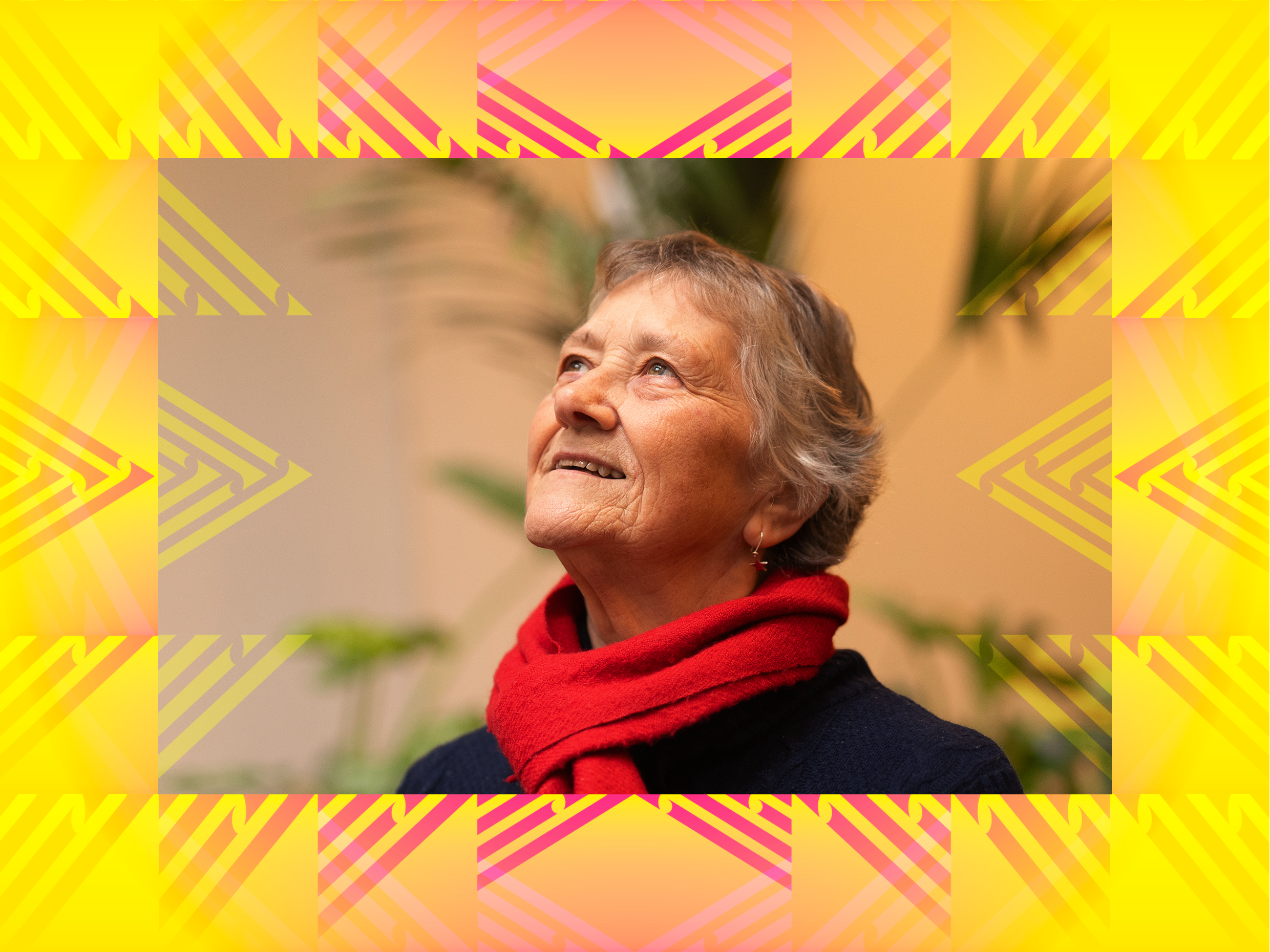 A picture of Jill from Innercity Wellington. She has short gray hair wearing a red scarf and a navy blue top, looking upwards with a slight smile, standing in front of a green plant in an indoor setting.