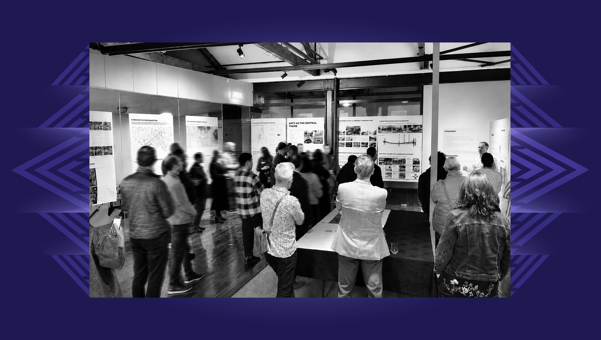 People gathered indoors at one of the early Te Aro Hā Courtenay workshops, looking at displays and posters on the walls outlining the vision and plan for the initiative. Some are conversing, and one person is taking a photo.
