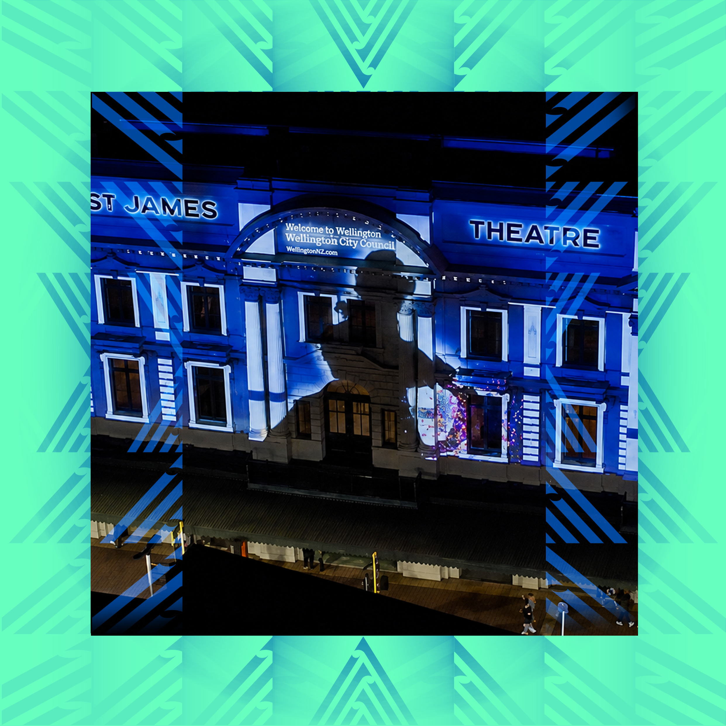 Night scene of the St James Theatre with neon lights. Signs read "St James Theatre" and "Welcome to Wellington" in blue lighting. The building has classical architecture with ornate details and large windows. A few people are walking nearby.