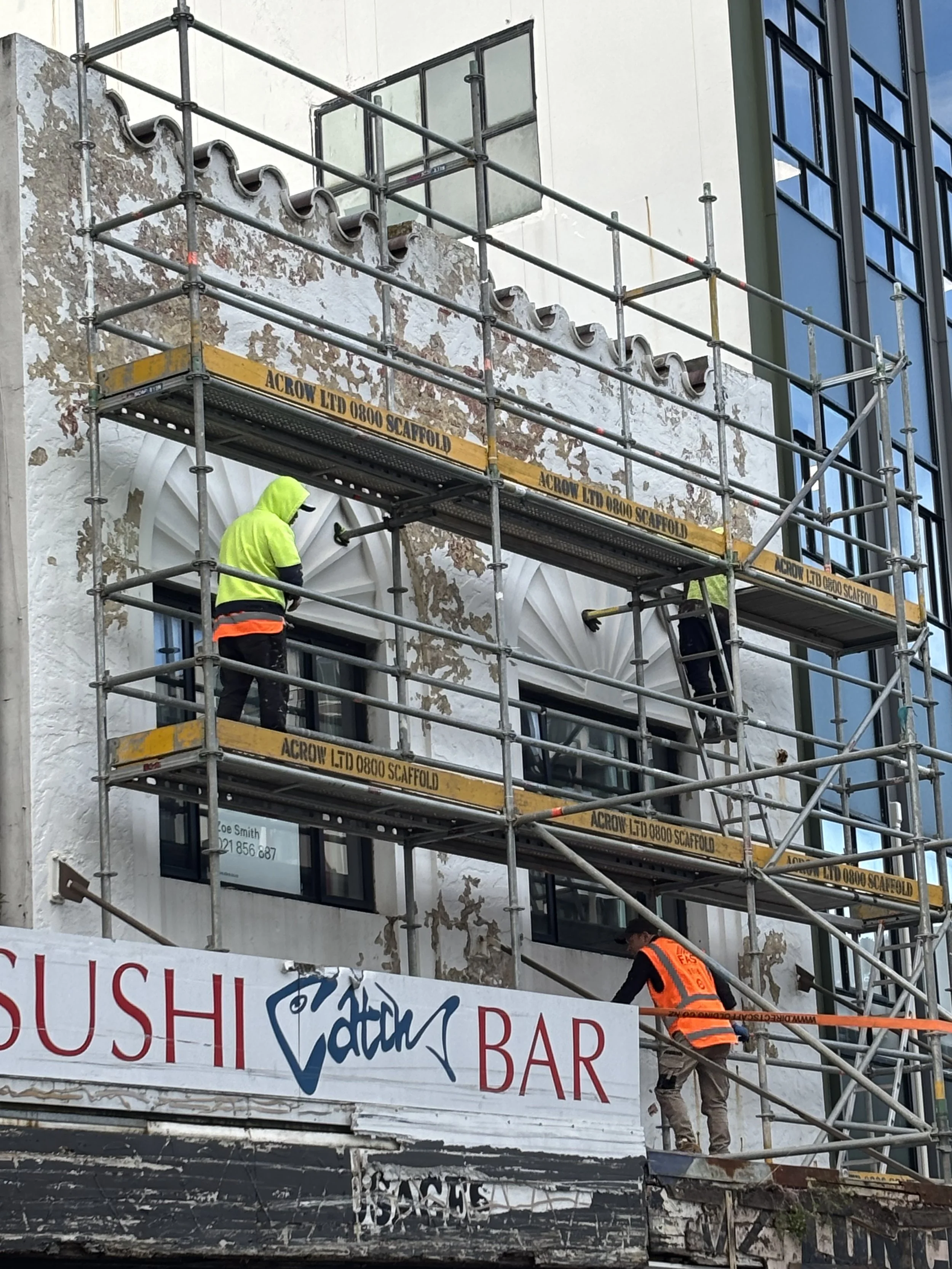 Construction workers in high-visibility vests working on a building facade with scaffolding and a partially painted wall. A sign for a sushi bar is visible at the bottom of the image.