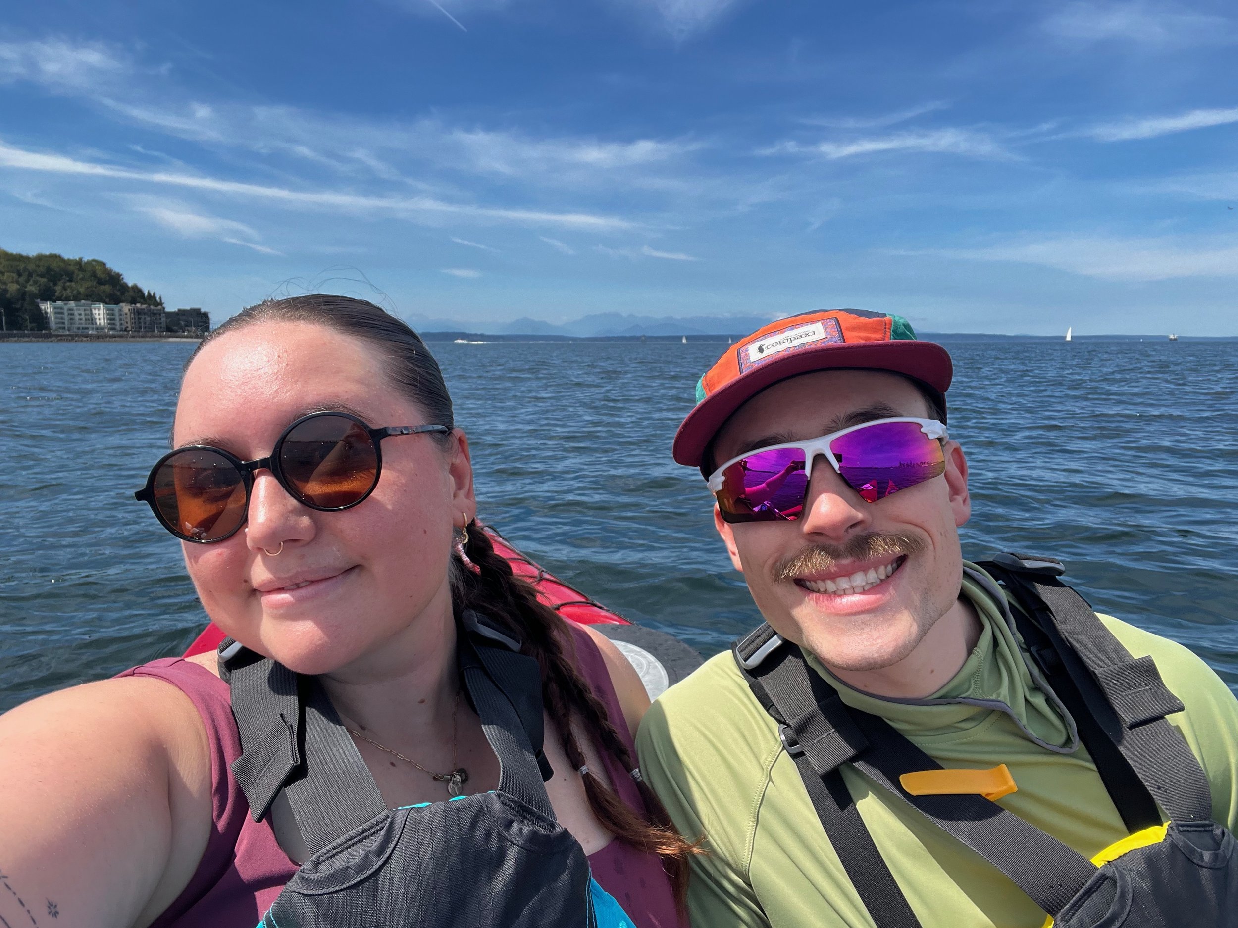 Sydney and Sean Healy smiling taking a selfie from their kayaks in the San Juans. The sky is blue with scattered clouds, and a shoreline and buildings visible in the background.