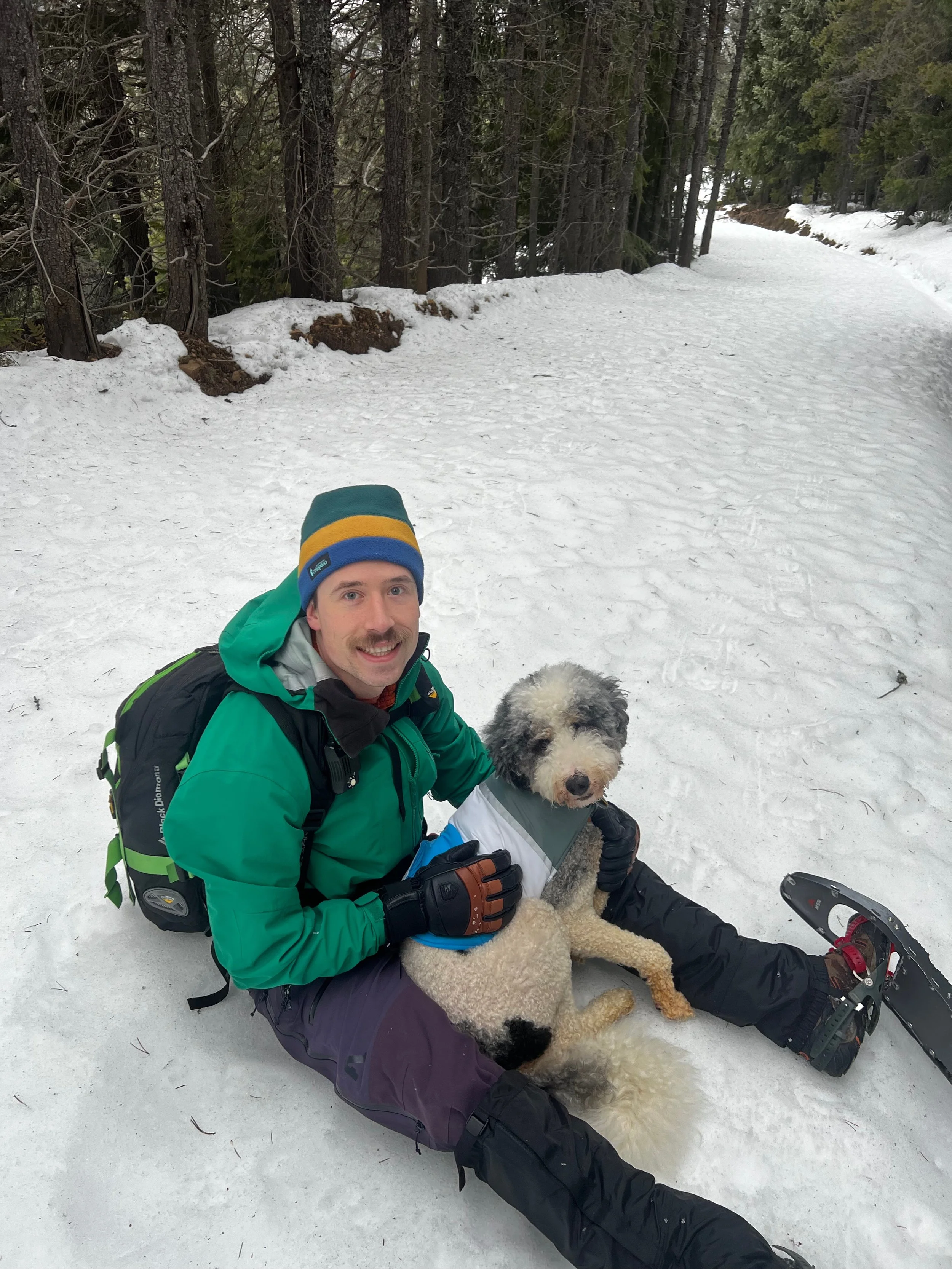 Sean and the dog, Ollie, smiling at the camera, sitting in the snow taking a break from snowshoeing in the Cascades.
