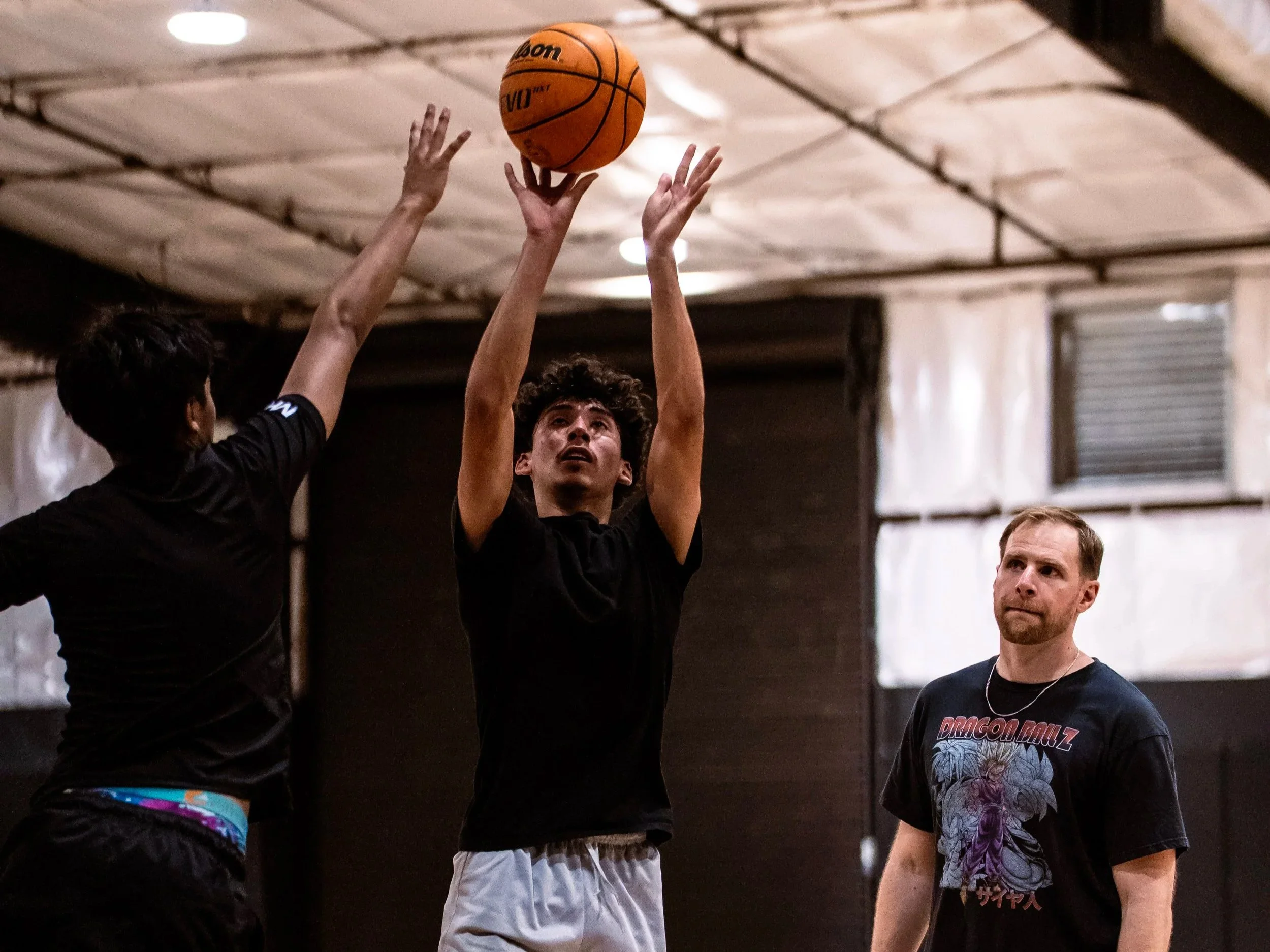 A basketball player in a black shirt is jumping to take a shot, with hands gripping a basketball above his head, while another player in black reaches with hands up, in an indoor gym.