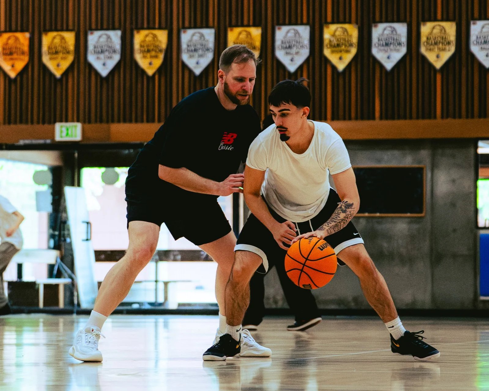 Two men are playing basketball in a gym. One is guarding the other, who is dribbling the ball. The man with the ball has tattoos on his arm and is wearing a white t-shirt and black shorts. The man guarding him is wearing a black t-shirt and black shorts.