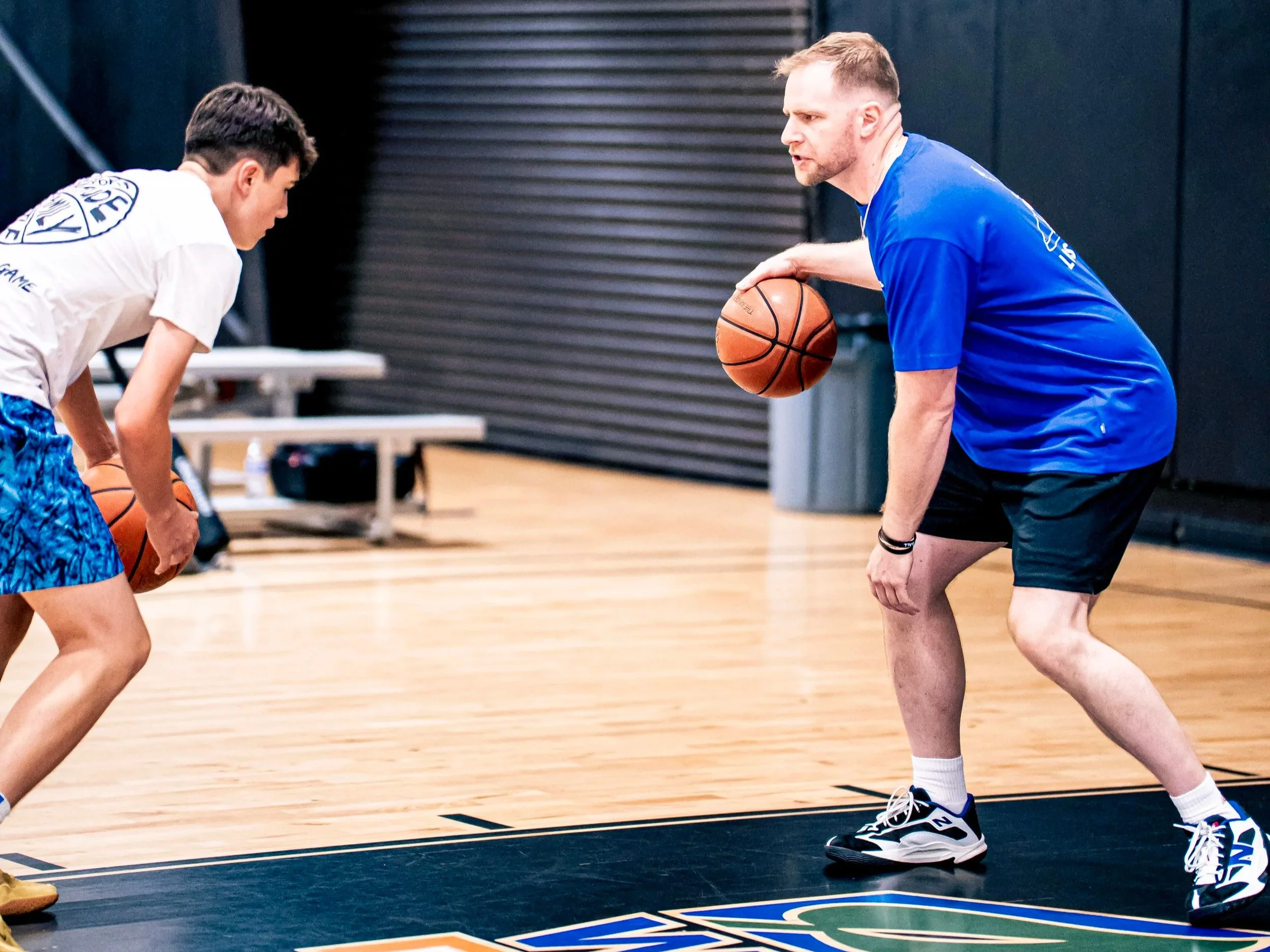 A man and a boy playing basketball indoors. The man is in a blue shirt and black shorts, holding a basketball, and the boy is in a white shirt and blue shorts. They are on a wooden court with black lines, and there are tables and black walls in the background.