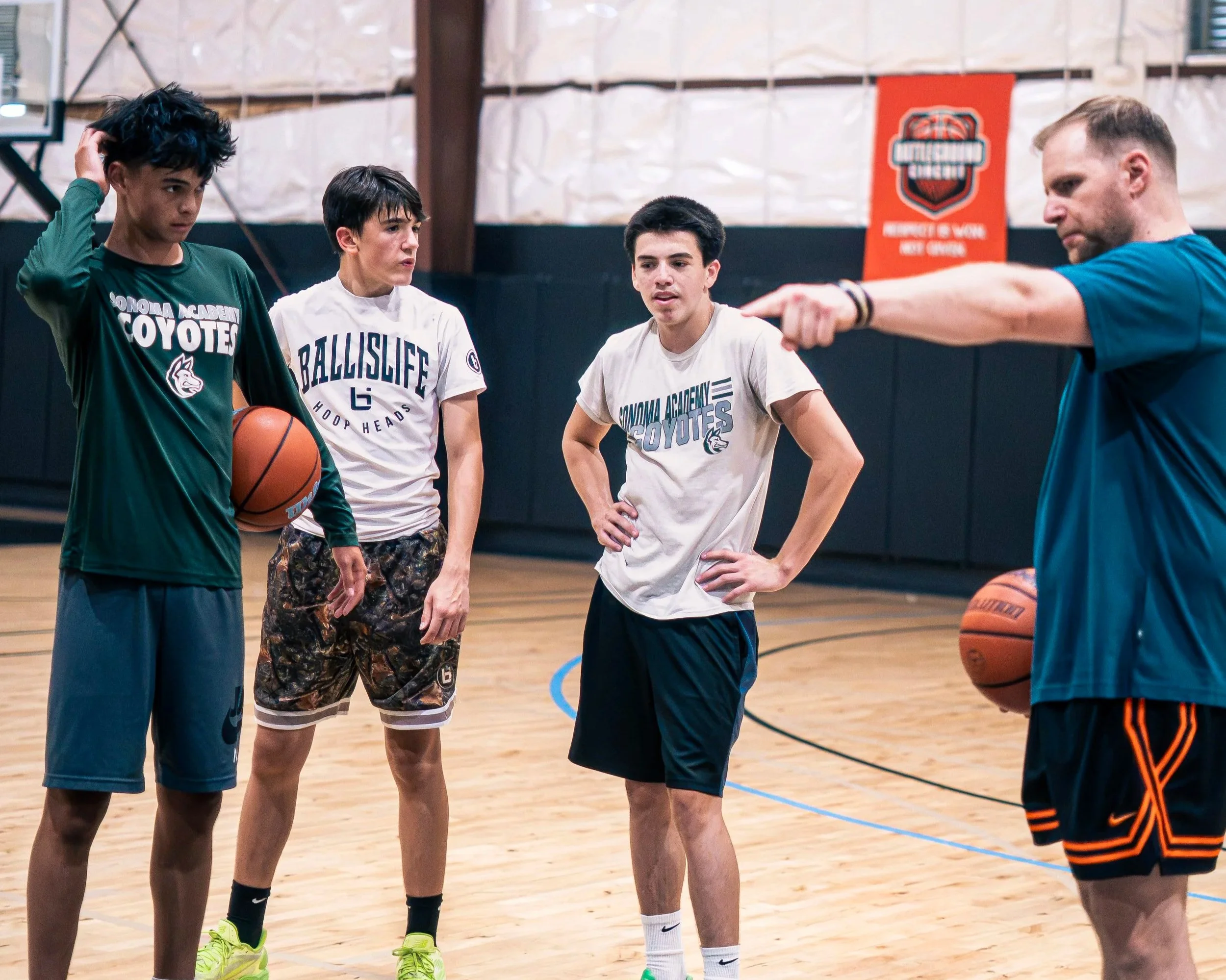 Group of young male basketball players standing in a gym, listening to their coach who is pointing and giving instructions. The players wear sports T-shirts and shorts, with one holding a basketball. The gym has a basketball court with a wooden floor and a basketball hoop visible in the background.