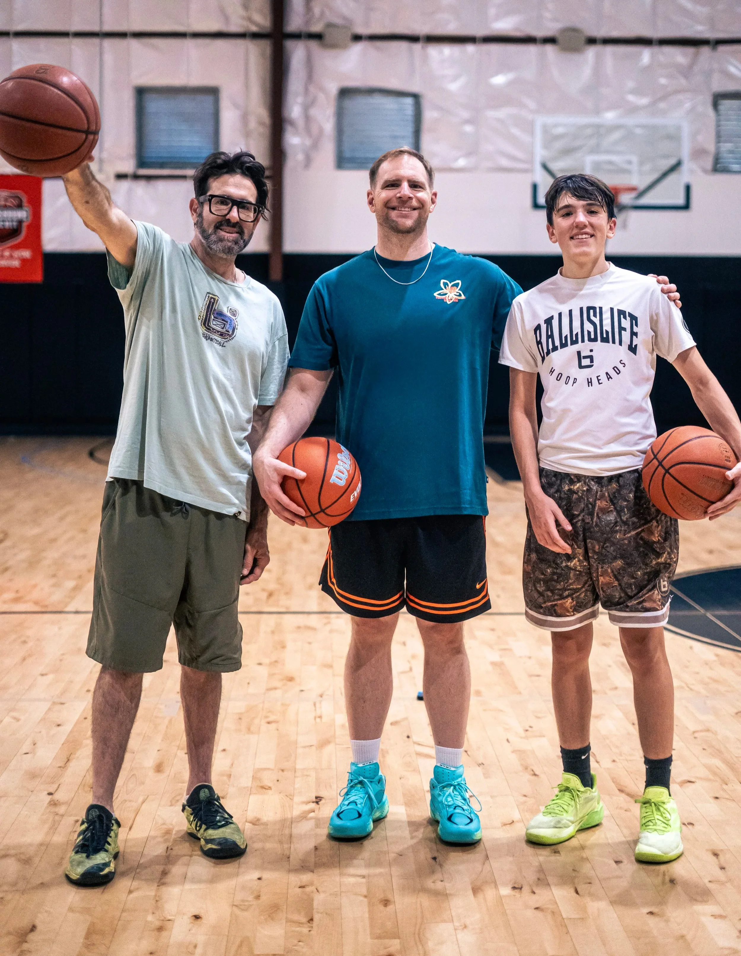 Three basketball players standing on an indoor court, smiling at the camera. The player on the left is holding a basketball, wearing glasses, a light-colored t-shirt, olive shorts, and yellow shoes. The player in the middle, also holding a basketball, is wearing a blue t-shirt, black shorts with orange accents, and bright blue shoes. The player on the right, holding a basketball, is wearing a white t-shirt with 'BALLISLIFE' printed on it, camouflage shorts, and neon green shoes.
