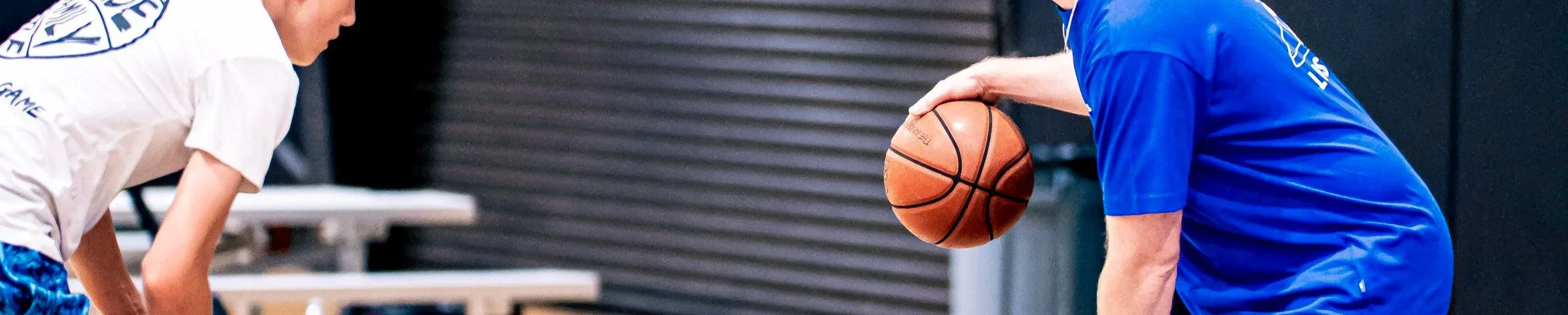 Two basketball players practicing in an indoor gym, one holding a basketball and the other bending over to receive the ball.