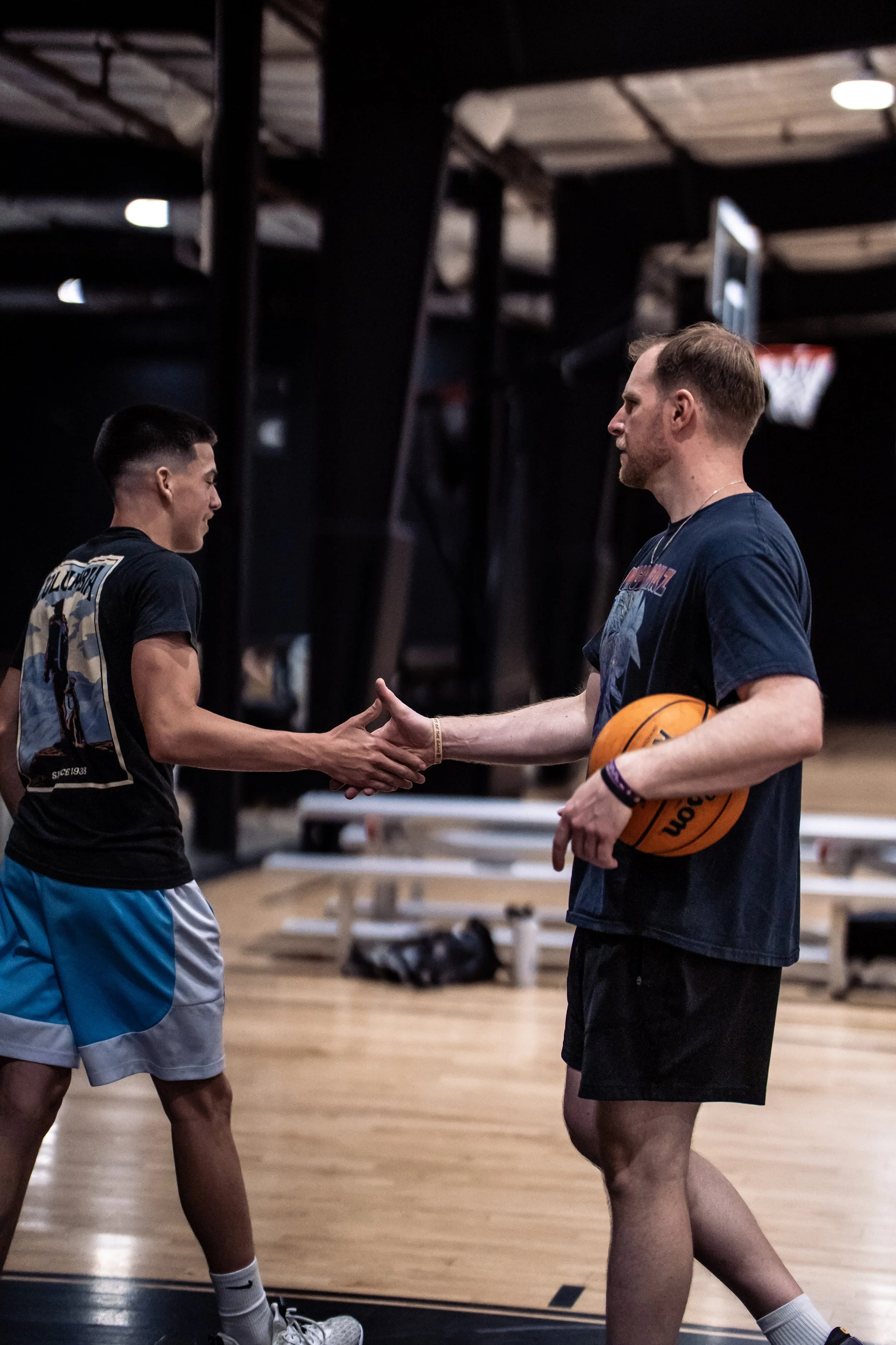 Two young men shake hands in a gym, one holding a basketball. They are standing on a basketball court, with gym equipment and a television in the background.
