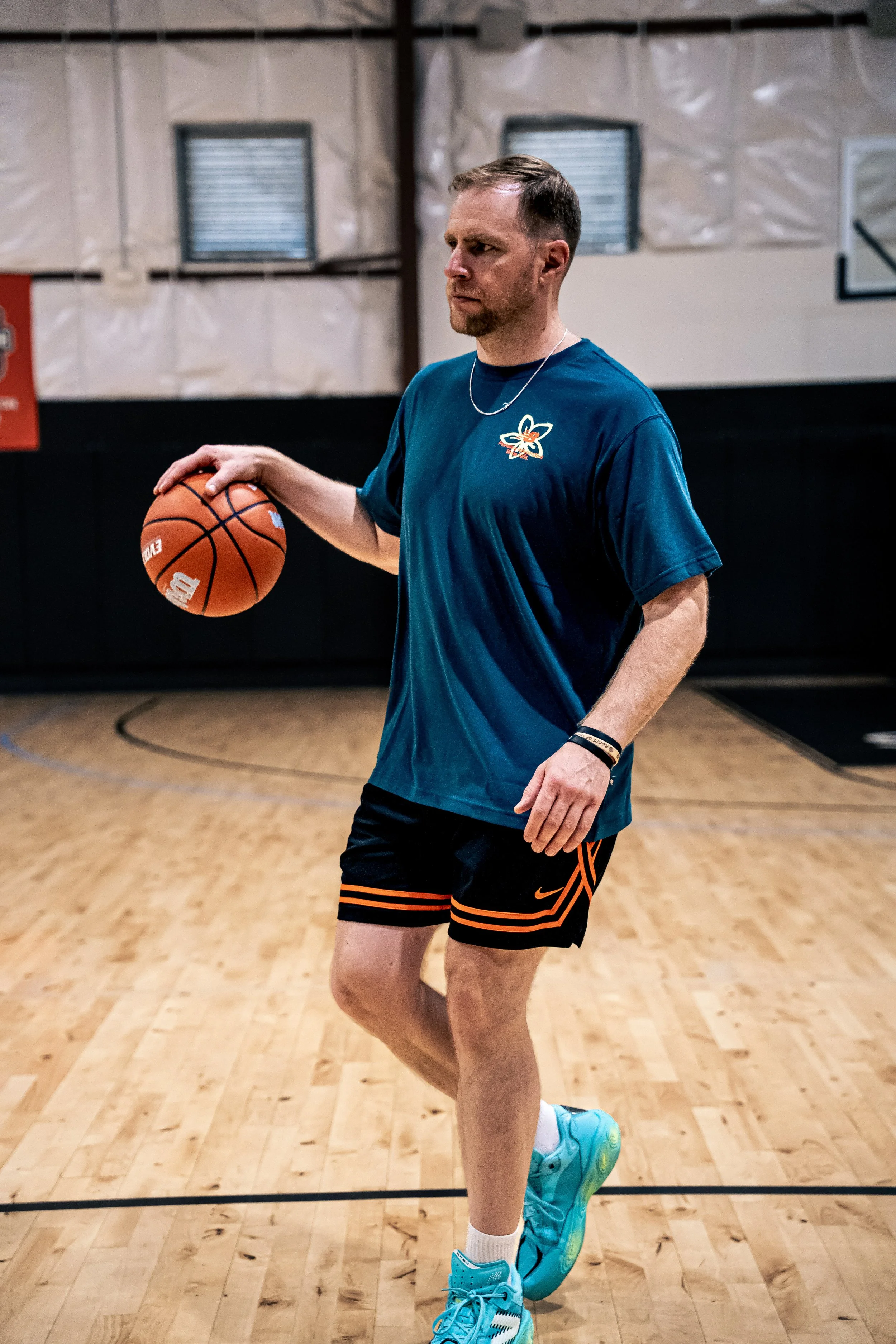A man is playing basketball on an indoor court. He is holding a basketball and appears to be preparing to shoot or pass. He wears a dark blue shirt with a flower logo, black shorts with orange stripes, and bright blue sneakers.