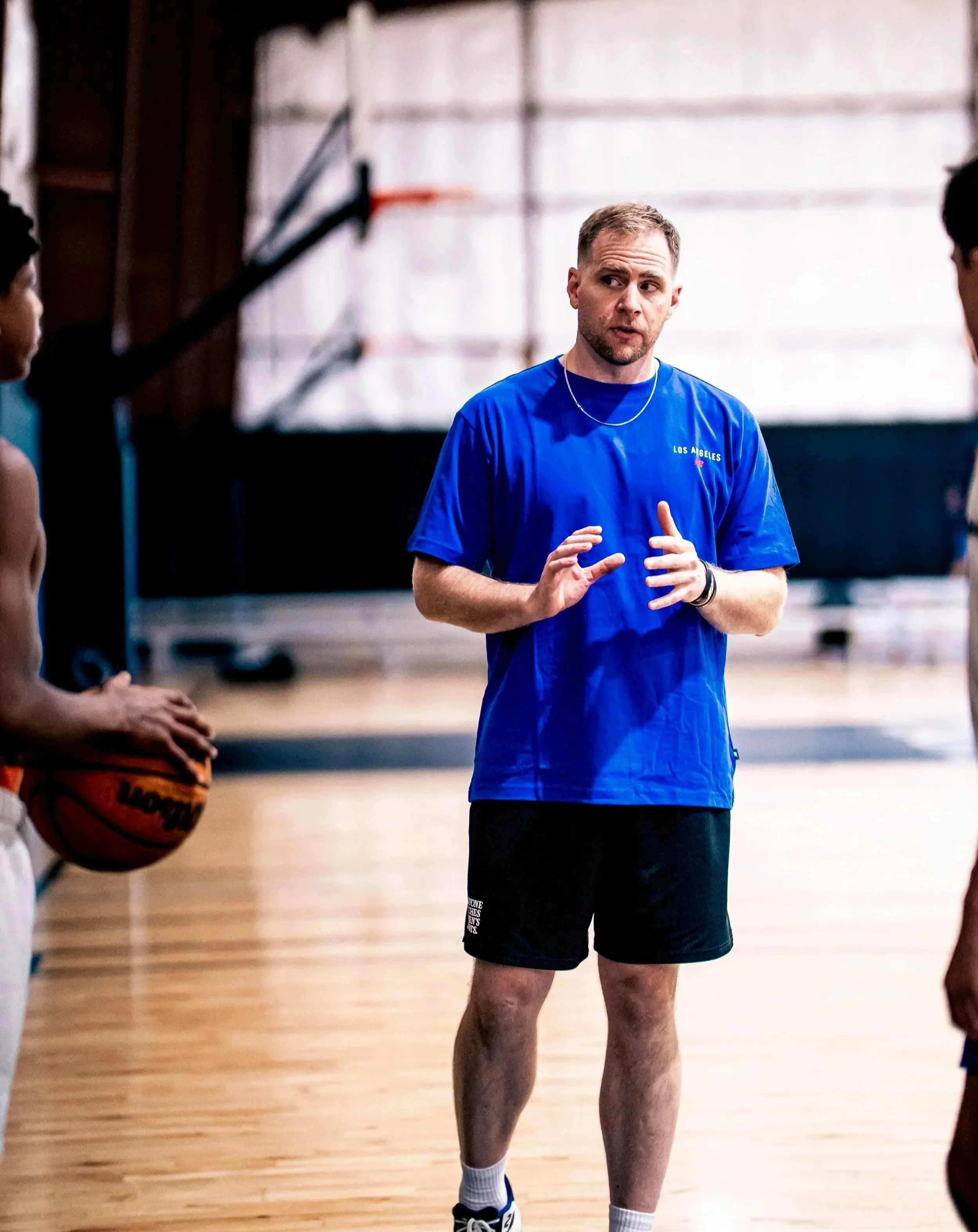 A man with short hair and a beard standing on a basketball court, talking to two other players, wearing a blue t-shirt with 'LOS ANGELES' written on it, black shorts, white ankle socks, and athletic shoes. There is a basketball hoop and backboard in the background.