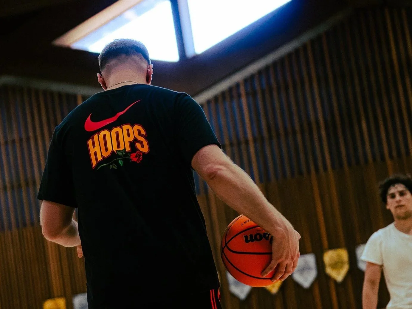 A person in a black t-shirt with the word "HOOPS" in orange and red, holding an orange basketball, inside a gym with wooden paneling walls and flags in the background.