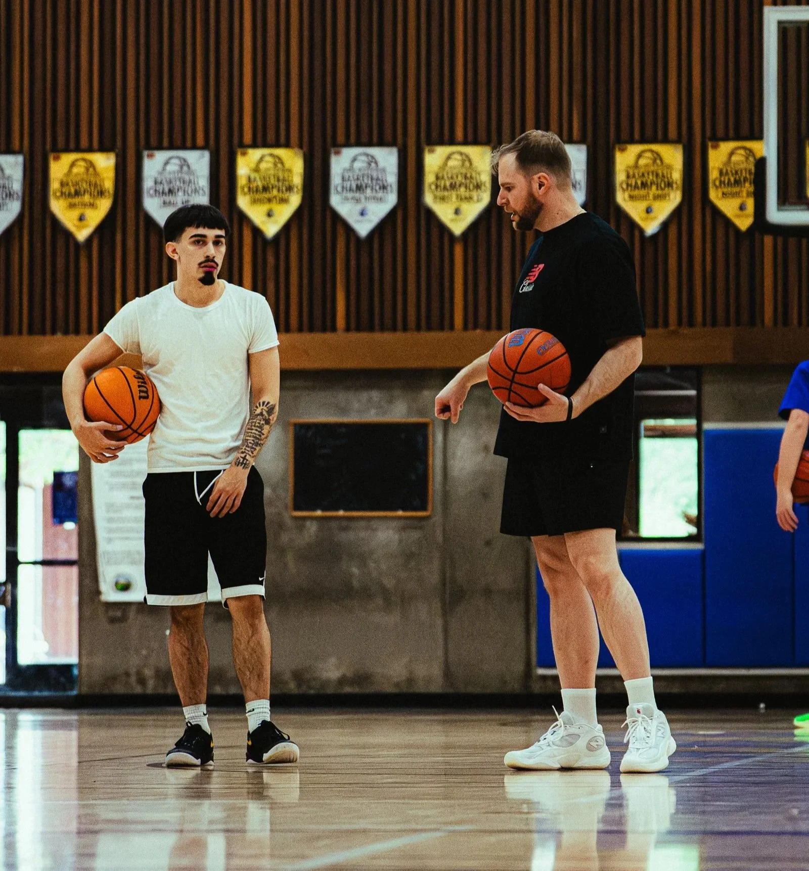 Two men standing on a basketball court in a gymnasium, each holding a basketball, engaging in a conversation. Background features a wooden wall with championship banners hanging.