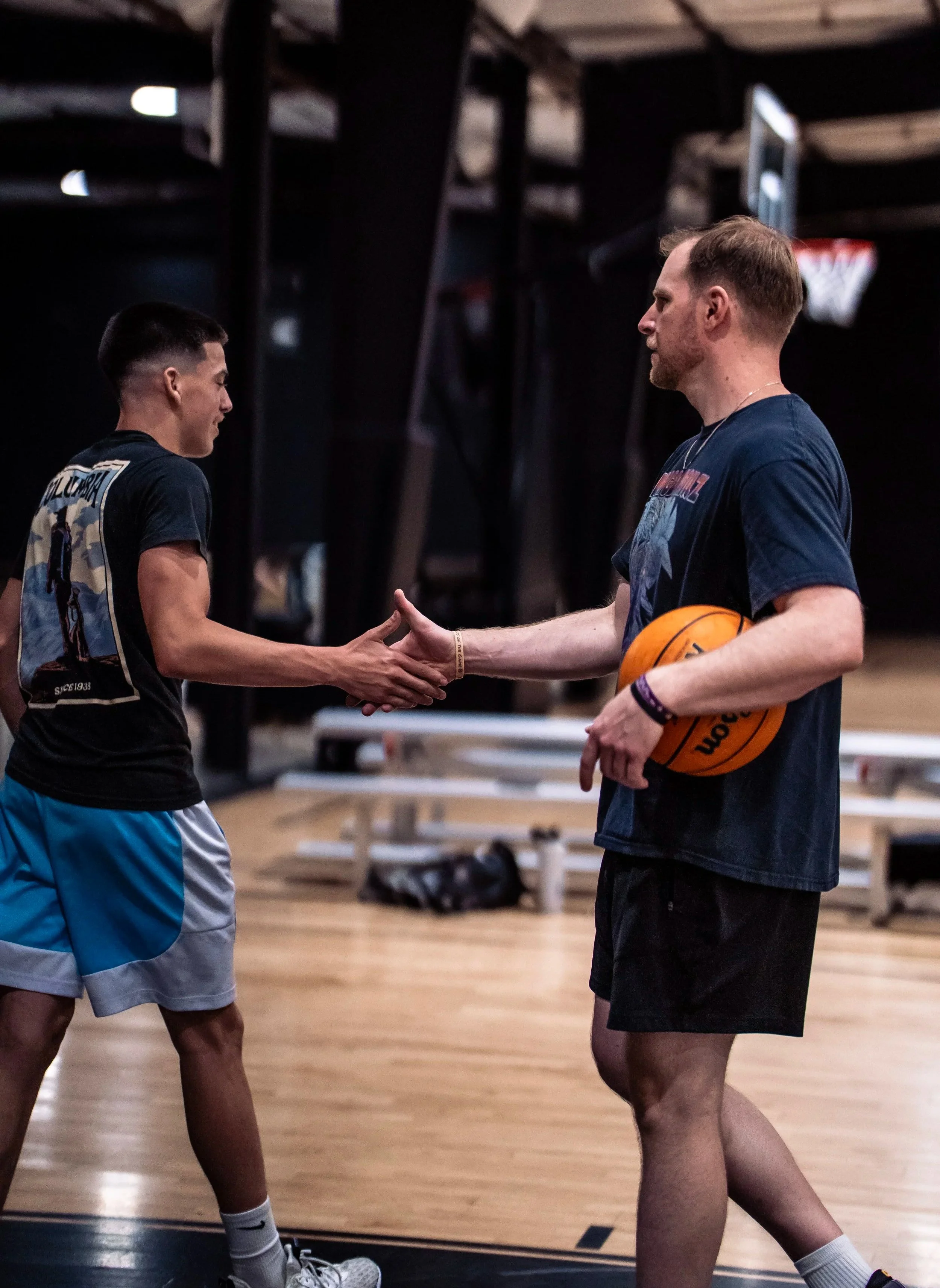 Two men shaking hands in a gym, one holding a basketball, with exercise equipment in the background.