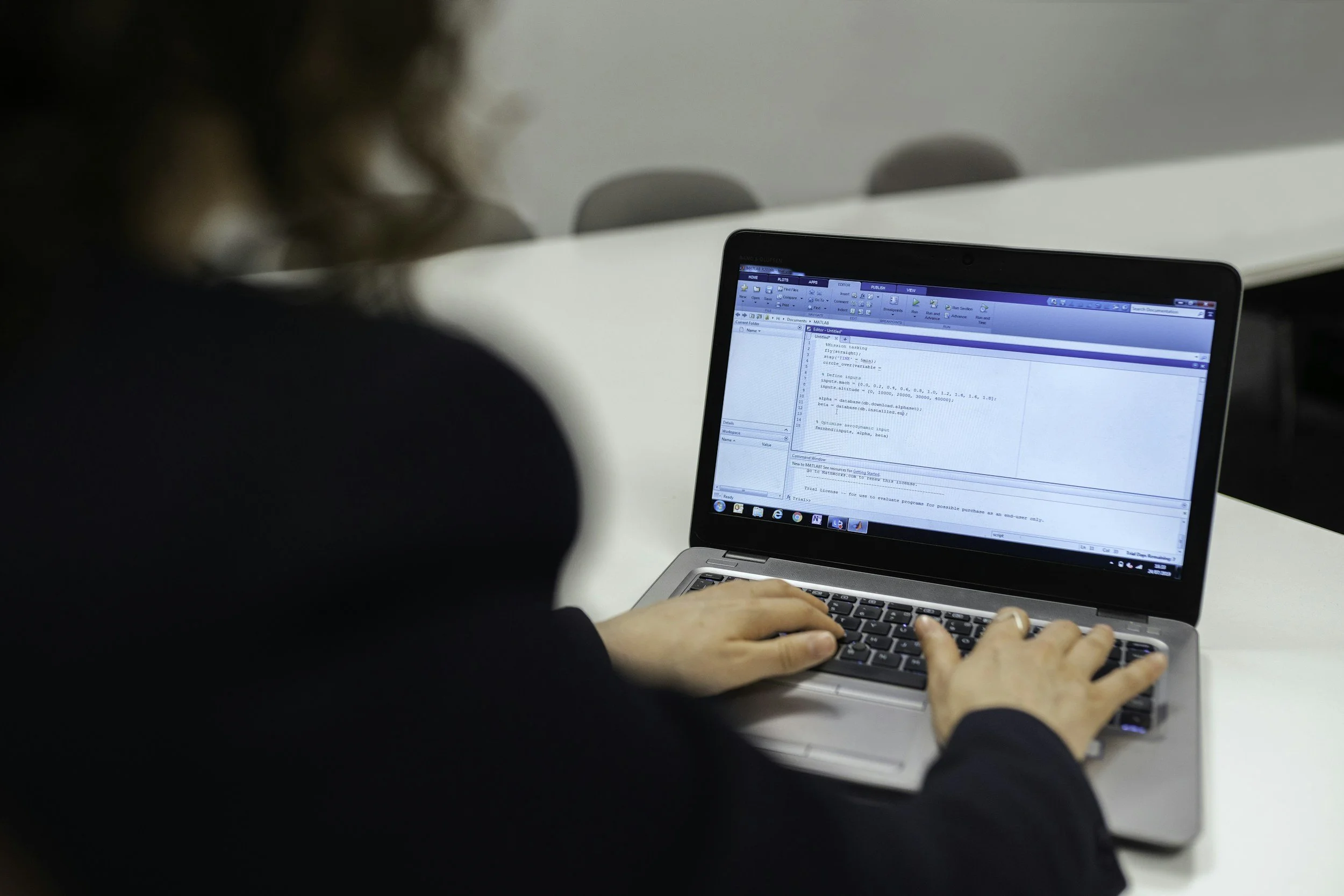Person using a laptop with programming code displayed on the screen in a room with chairs in the background.
