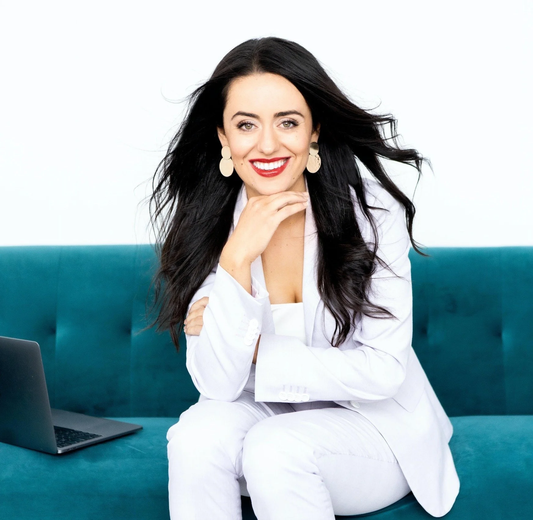 A woman with long black hair, light skin, and red lipstick smiling while sitting on a teal couch. She wears large white earrings, a white blazer, and white pants. A laptop is open beside her.