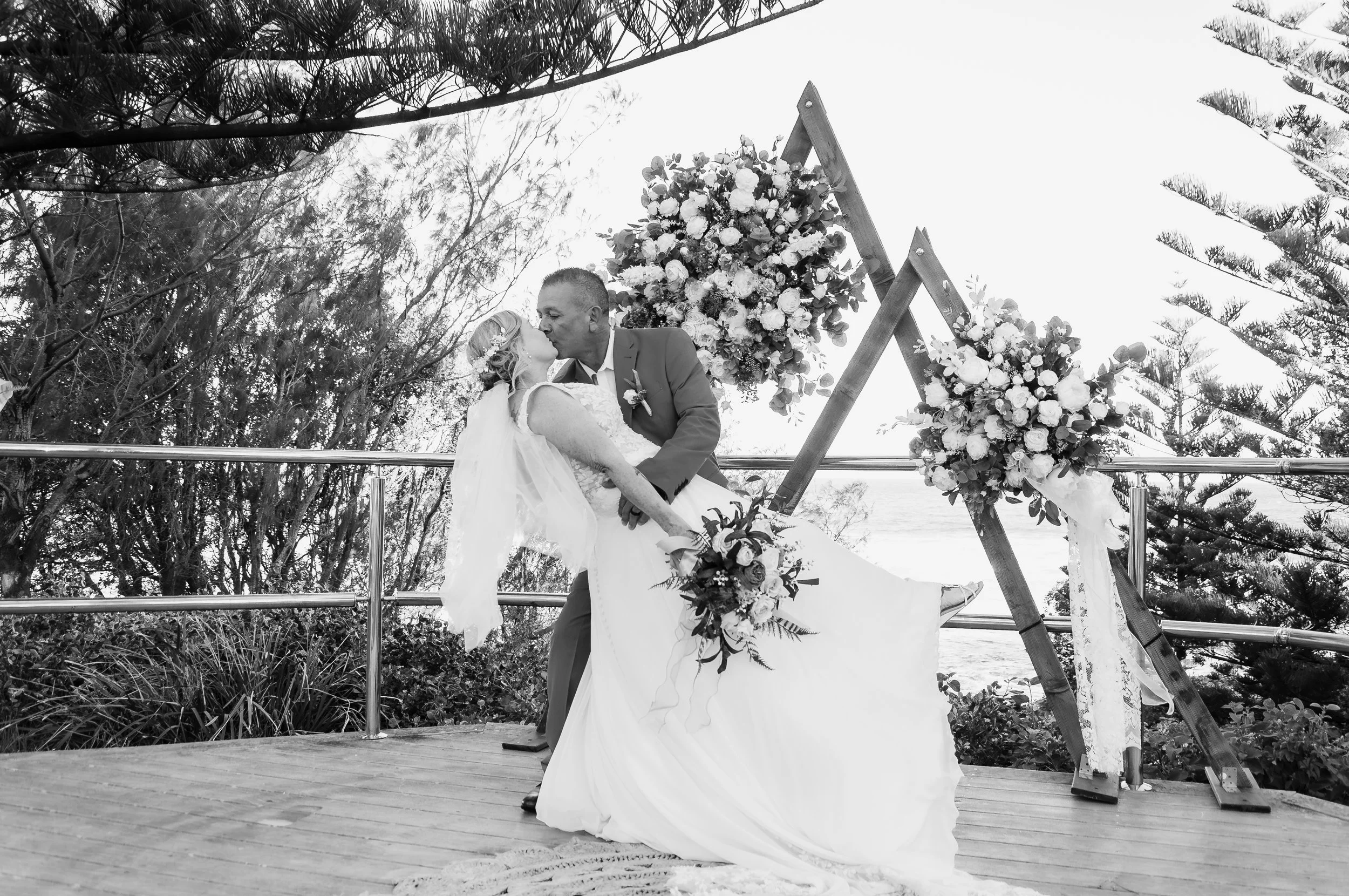 A bride and groom kissing during their outdoor wedding ceremony, with a floral arch and wooden structure behind them, surrounded by trees.