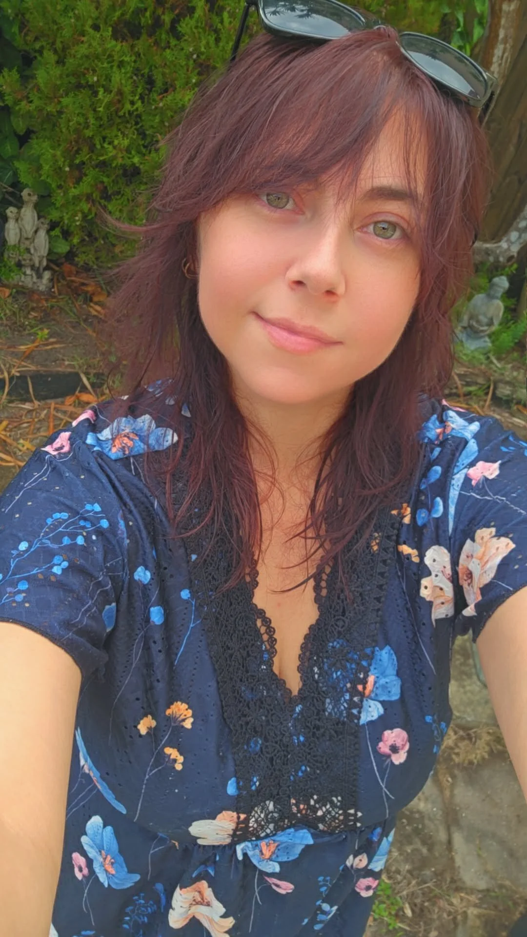 A woman with red-brown hair and green eyes taking a selfie outdoors, wearing glasses on her head and a dark blue floral dress.