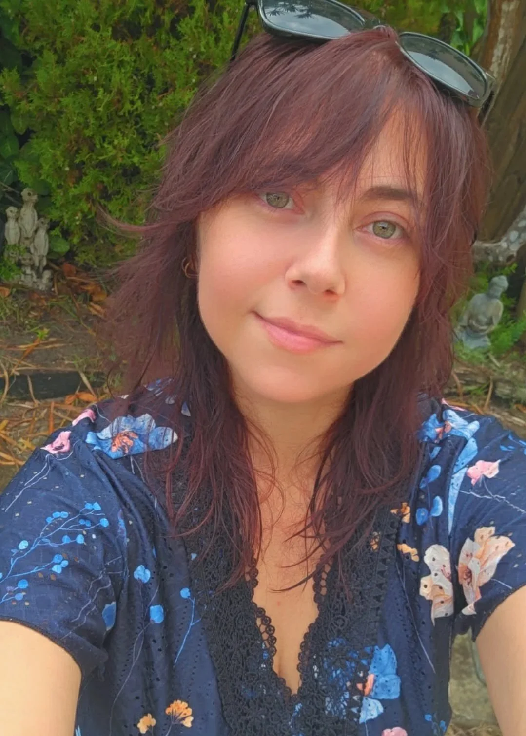 A woman with reddish-brown hair and sunglasses on her head taking a selfie outdoors, with greenery and small garden statues in the background.