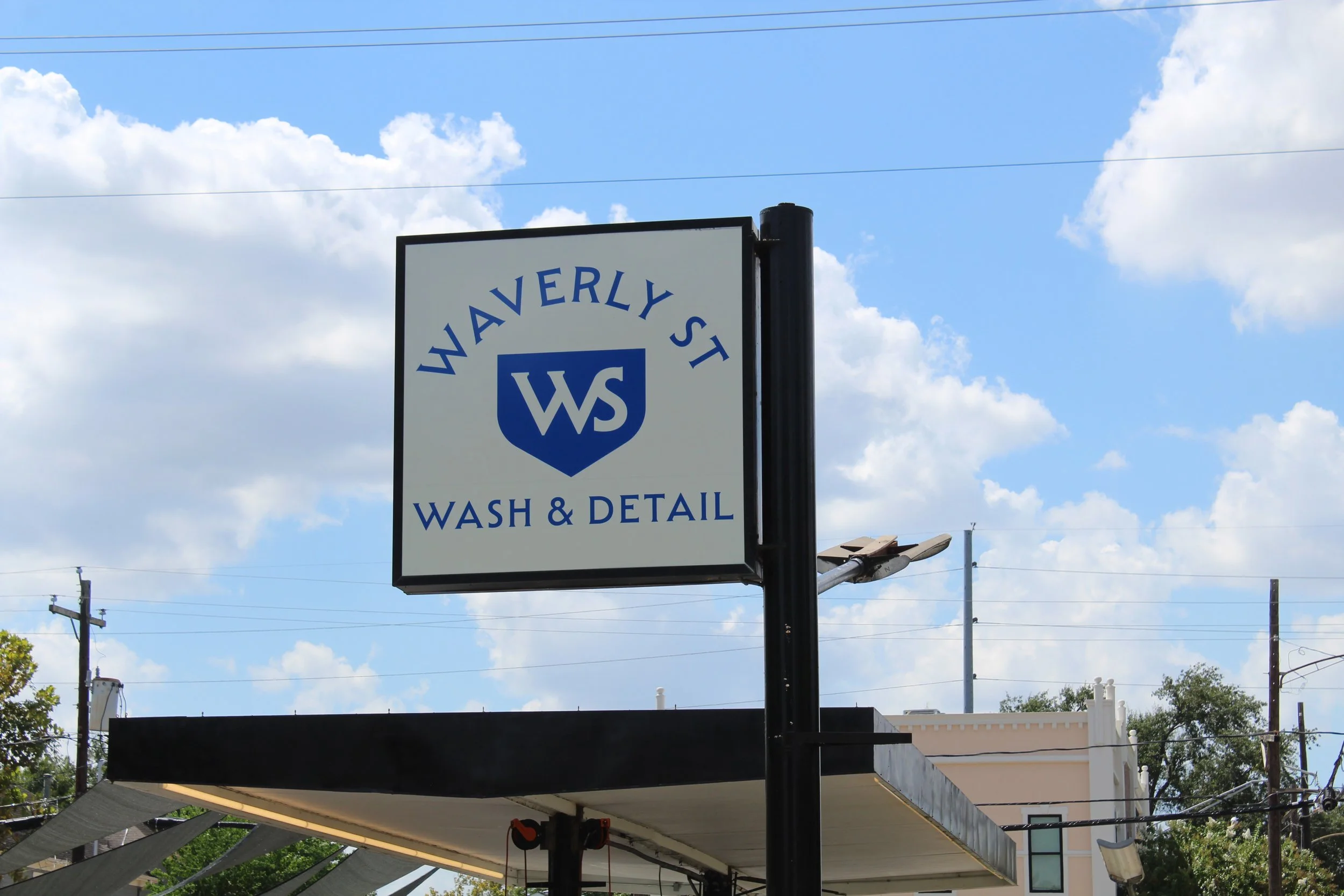 An outdoor sign reads 'Waverly St Wash & Detail' against a backdrop of a blue sky with clouds, power lines, and nearby rooftops.