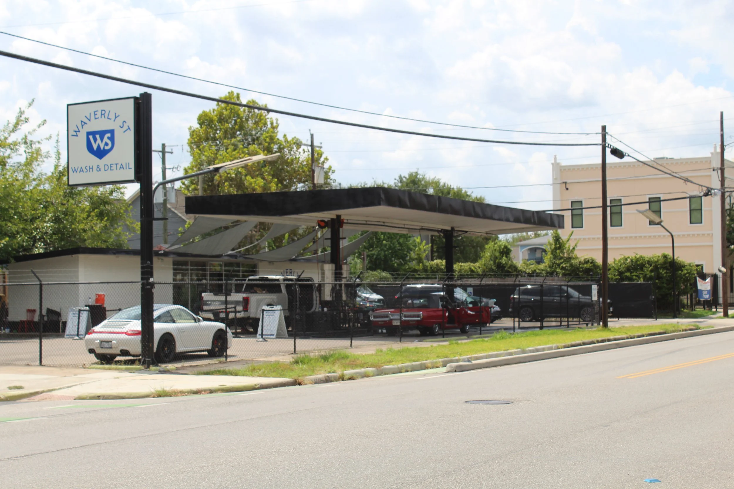 Street view showing a parking lot with several cars, a business sign for 'Waverly St Wash & Detail,' trees, utility poles, and buildings under a partly cloudy sky.