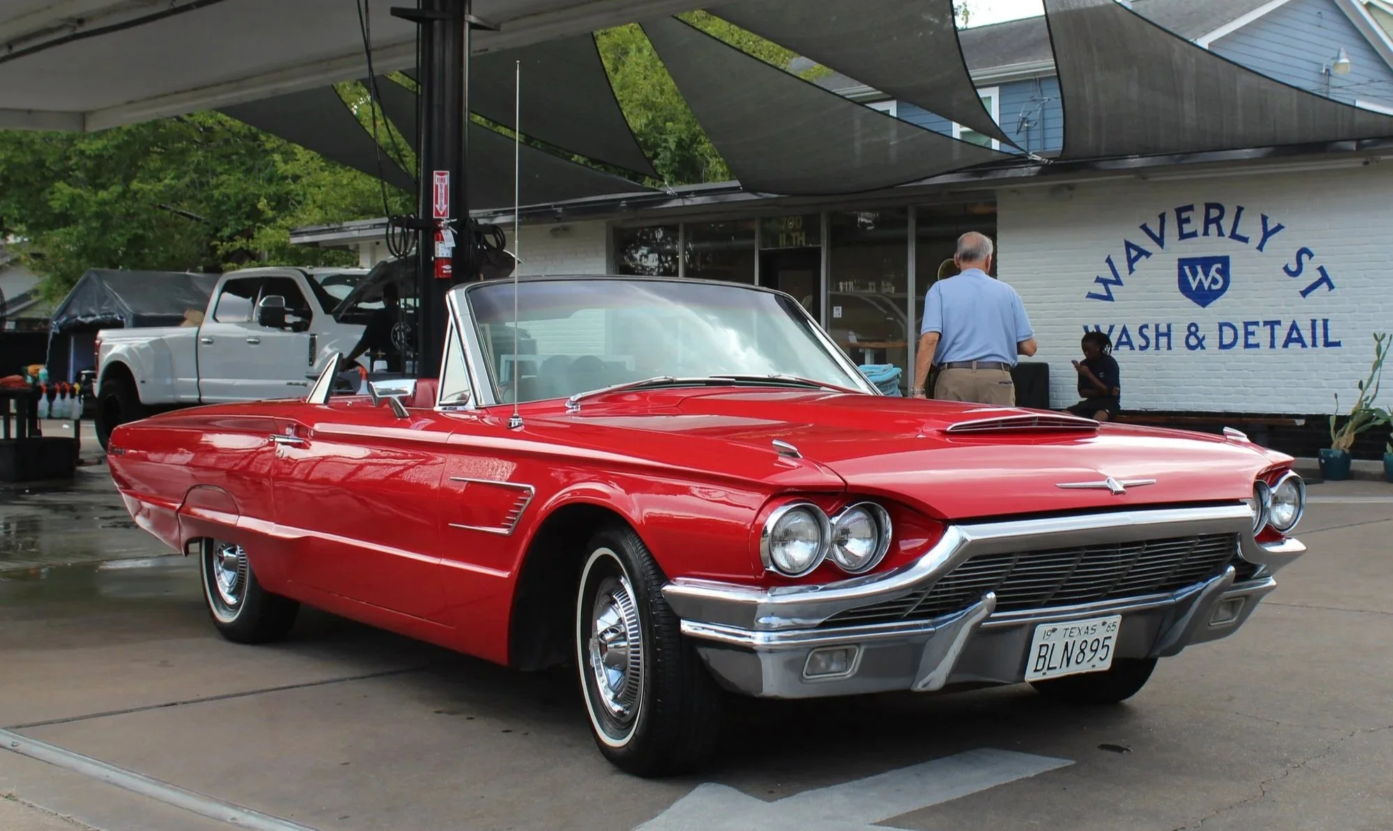 Red vintage convertible car parked outside a car wash and detail shop with a white pickup truck in the background.