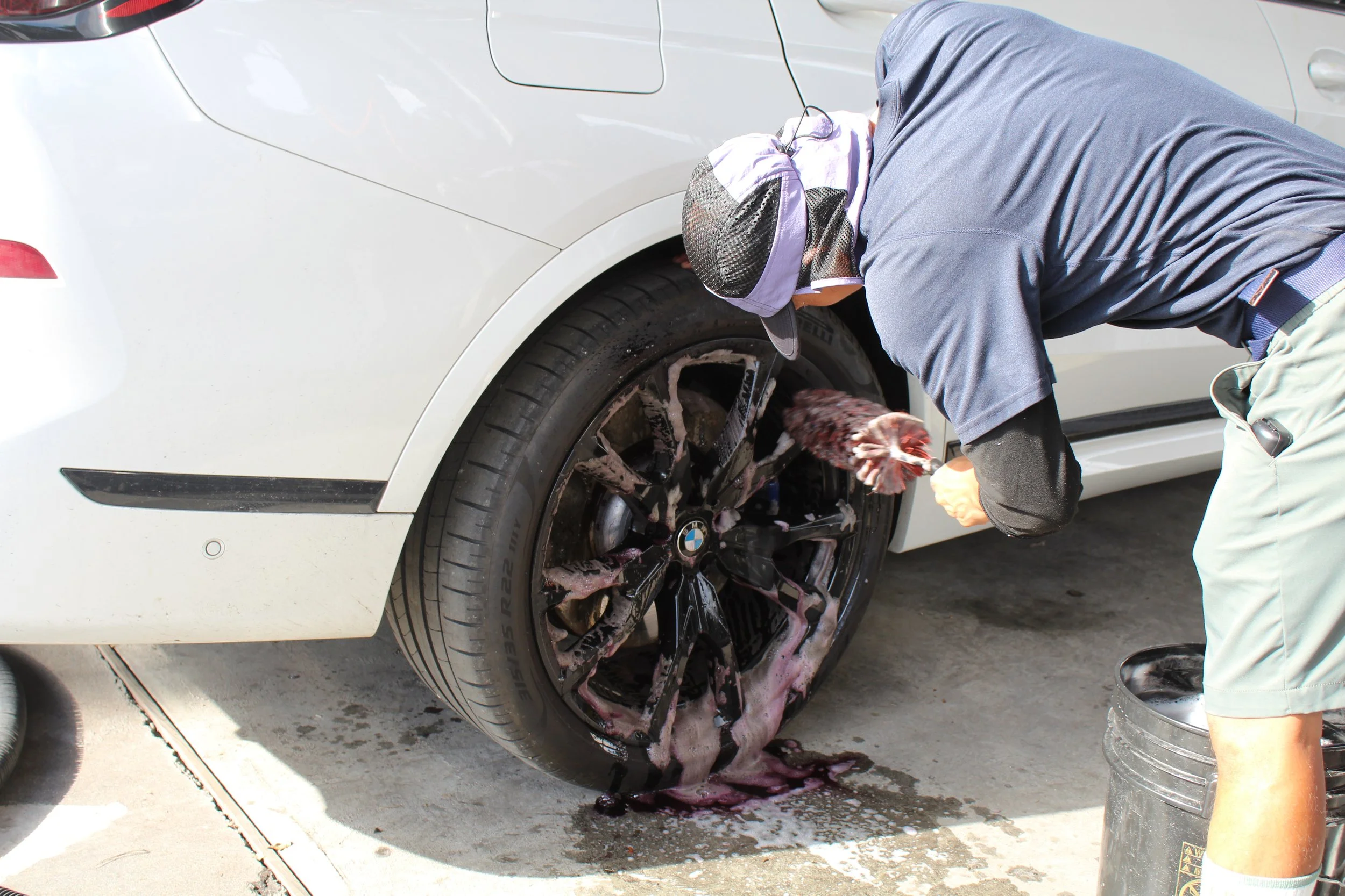 Person cleaning the black alloy wheel of a white car with soap suds spilled on the ground.