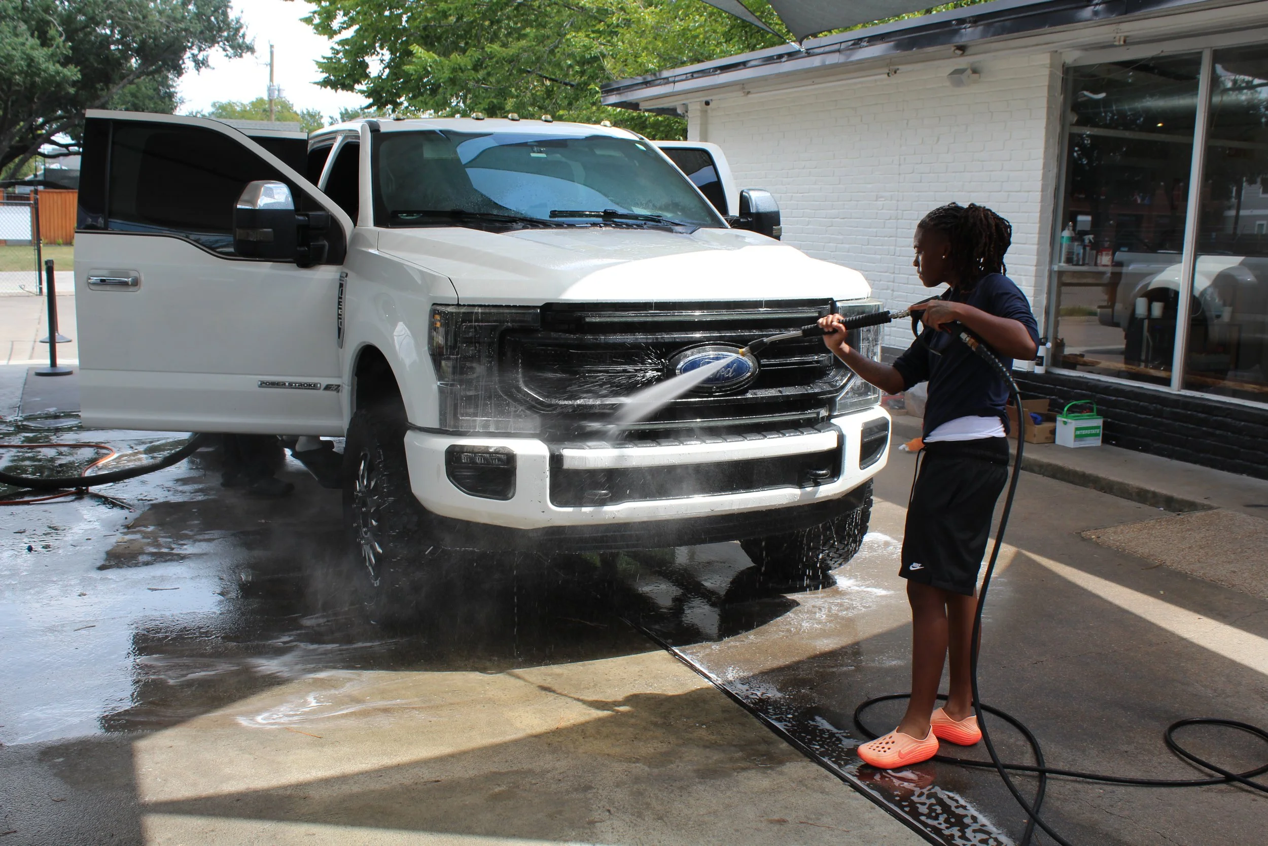 A person washing a white pickup truck with a long hose and water spray outside a building with a brick wall and a large window.