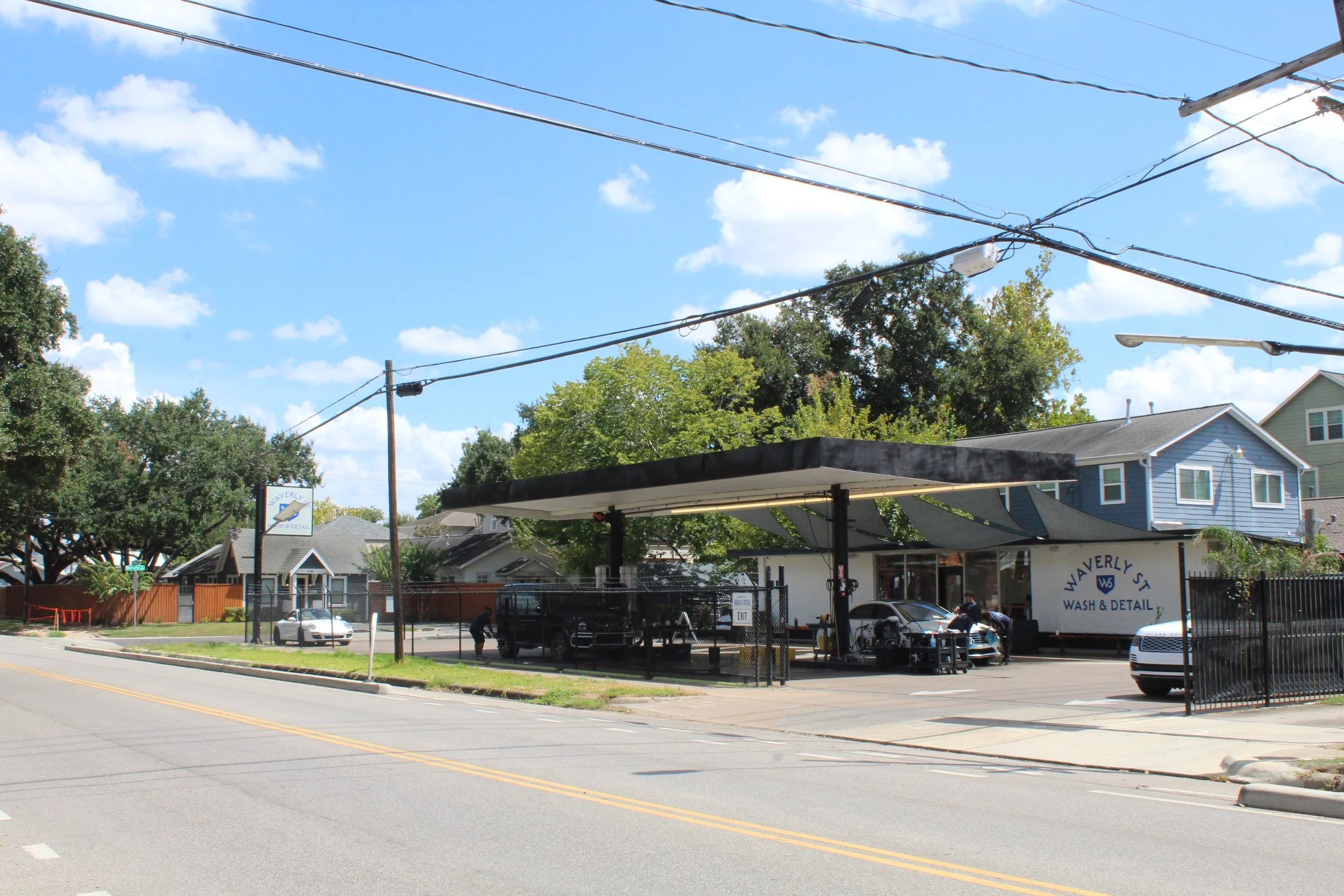 Street view of a commercial area with a car wash business, cars parked and being serviced, a few workers, electric wires overhead, residential houses in the background, and a bright blue sky with some clouds.