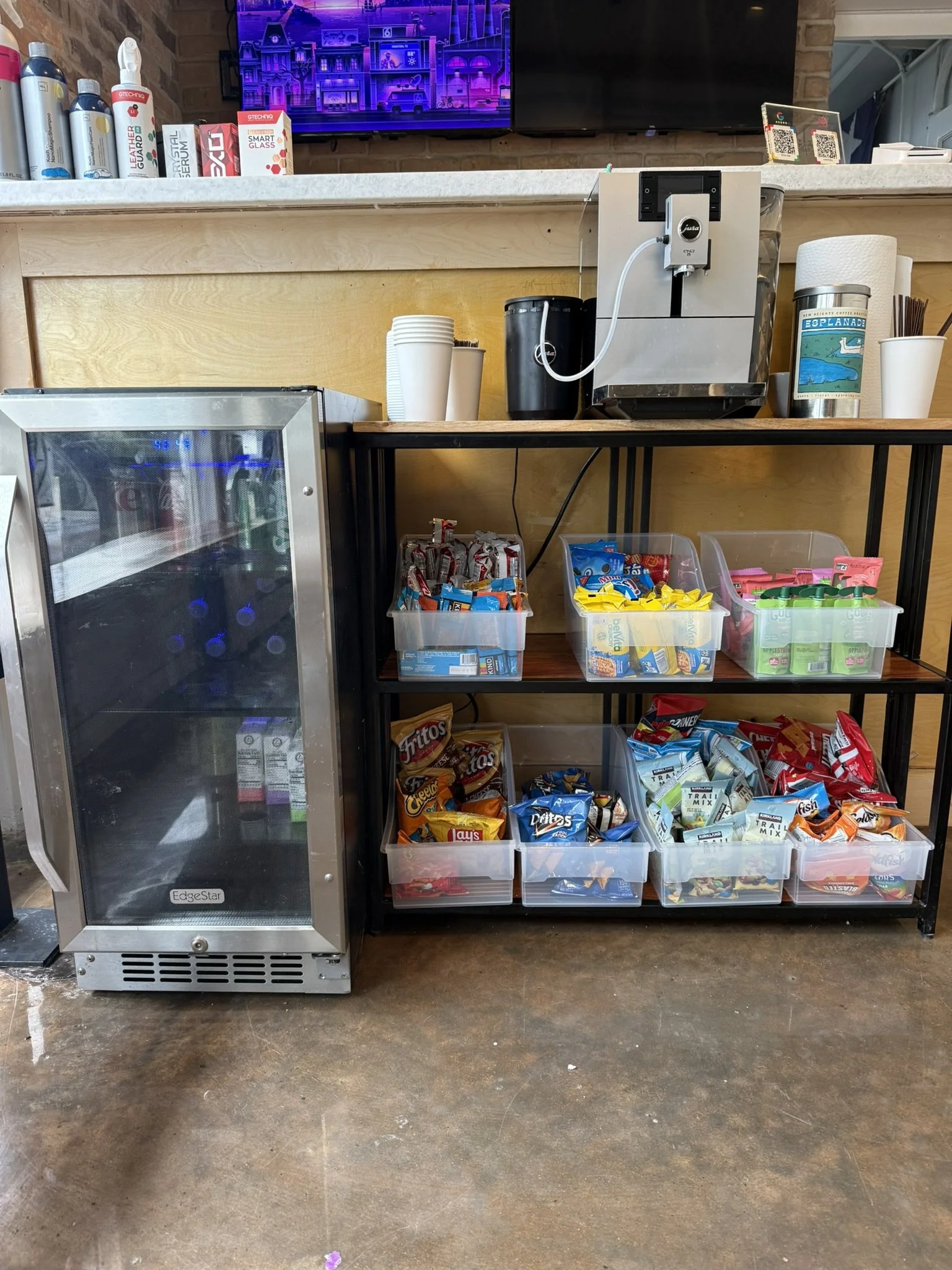 A snack station with a beverage cooler, snack containers filled with various chips and snacks, a coffee machine, cups, and a water bottle, situated on a wooden and metal table against a brick wall.