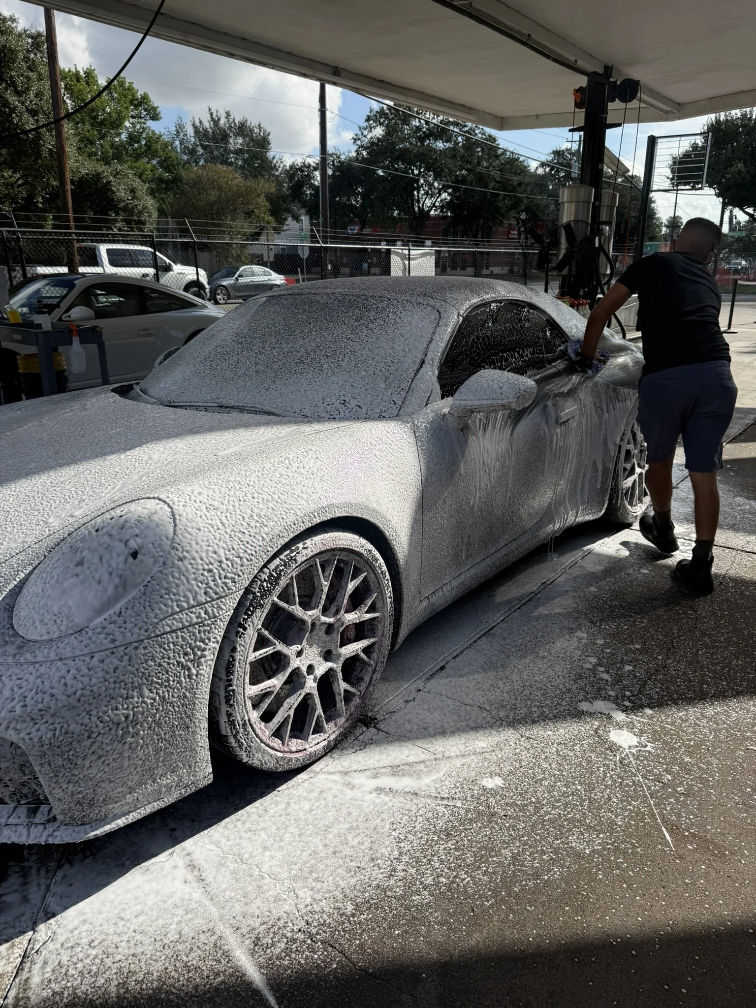 A man washing a silver sports car with foam at a car wash station. The car is covered in soap suds, and another white car is visible in the background. There are trees and other vehicles outside, and the car wash is in an outdoor area with a roof overhead.