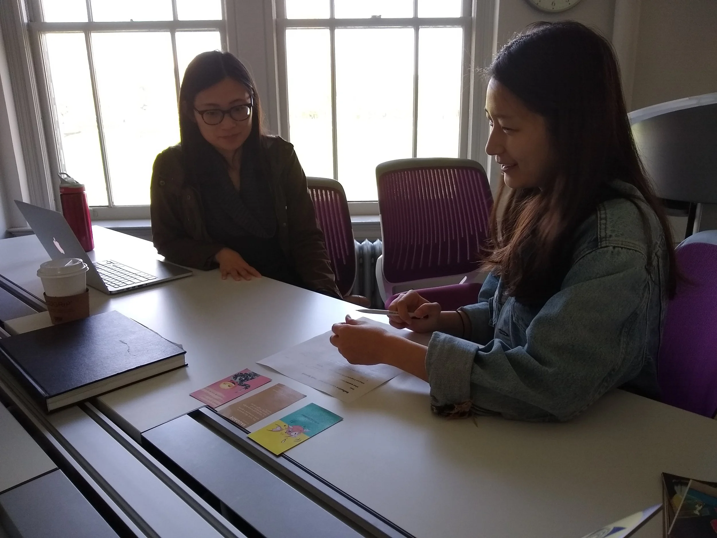 Two women sitting at a table in a room with large windows. One woman has dark hair and glasses, the other has long dark hair. The table has books, pamphlets, a laptop, a coffee cup, and a pink water bottle.