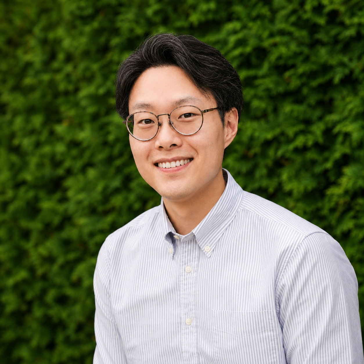 Portrait of a young Asian man with black hair and glasses, wearing a light striped button-up shirt, smiling in front of a lush green hedgerow.
