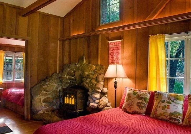 Cozy bedroom with wooden walls, a stone fireplace, and a red bed with floral pillows. Yellow curtains frame a window overlooking trees, and a floor lamp stands beside the bed.