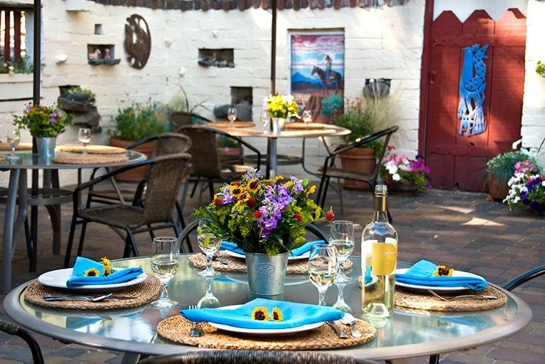 Outdoor dining table set with four plates, blue napkins, wine glasses, a bottle of white wine, and a flower centerpiece in a decorated patio area.