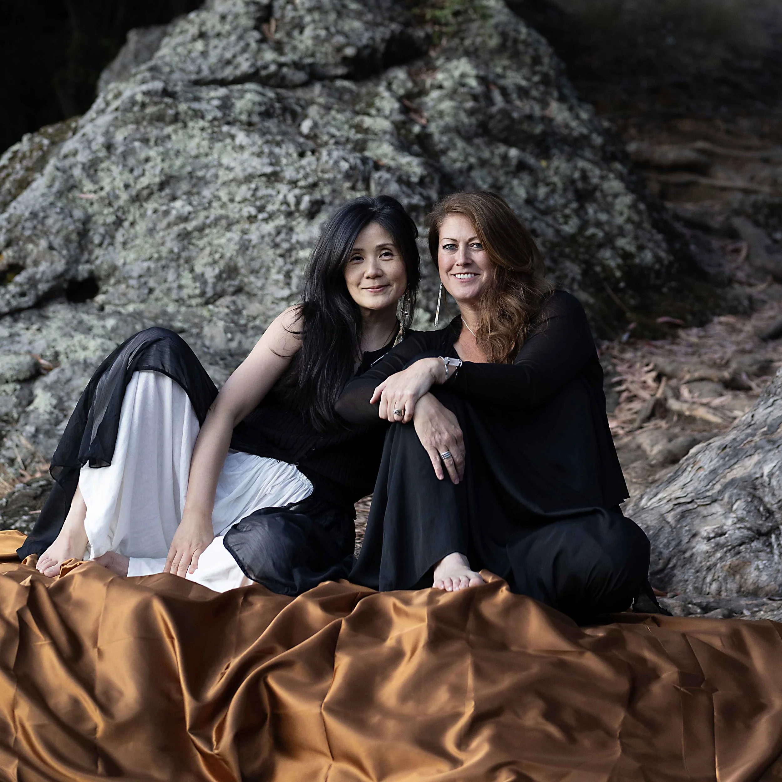 Two women sitting on the ground outdoors in front of a large rock formation, smiling at the camera. One woman has long black hair and the other has long brown hair. They are dressed casually with the woman on the left wearing a white skirt and the woman on the right in black clothing.