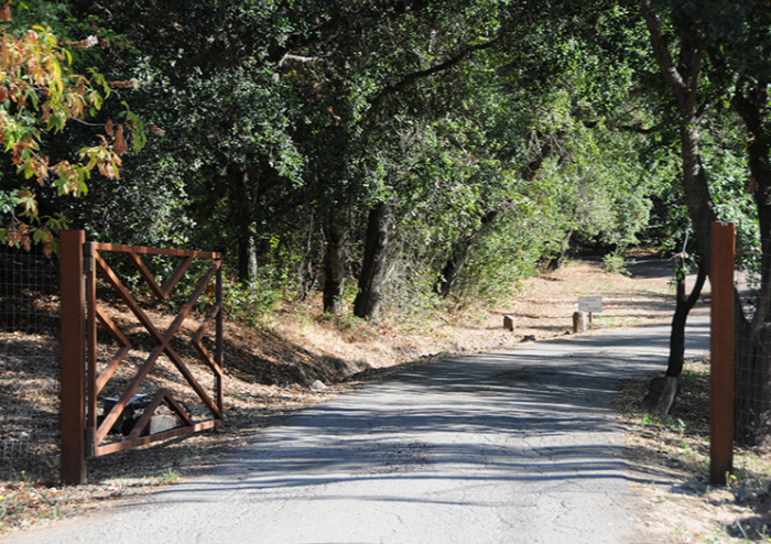 A dirt trail leading into a wooded area, with a partially open wooden gate on the left side and a fence on the right side, surrounded by green trees and bushes.