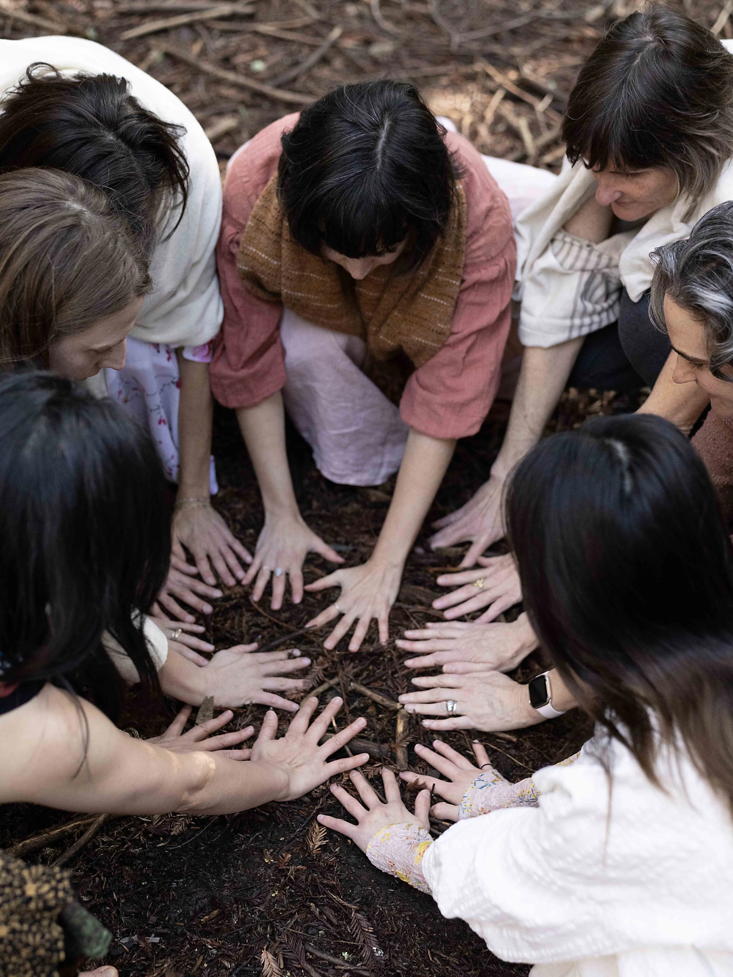 Group of people kneeling on the ground in a circle, with their hands touching dirt or soil, possibly planting or working on something together outside on a forest floor covered with leaves and twigs.
