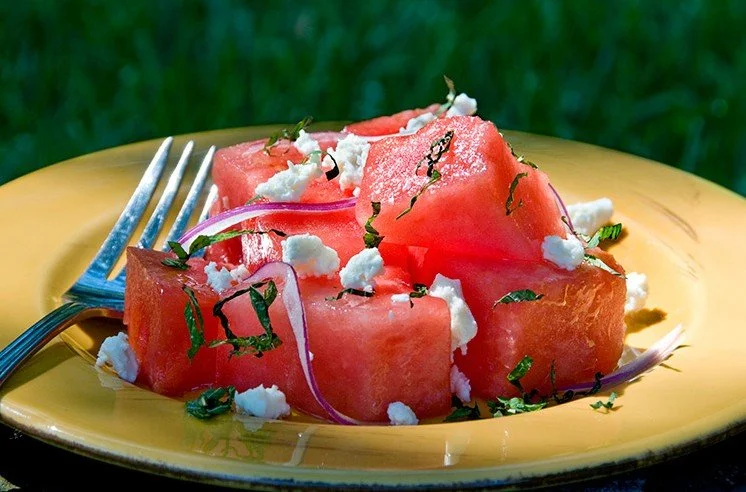 Slices of watermelon topped with crumbled cheese, herbs, and red onion on a yellow plate with a fork