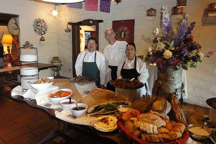Three chefs standing behind a table filled with various dishes, breads, and condiments in a warmly decorated restaurant with flowers and artwork.