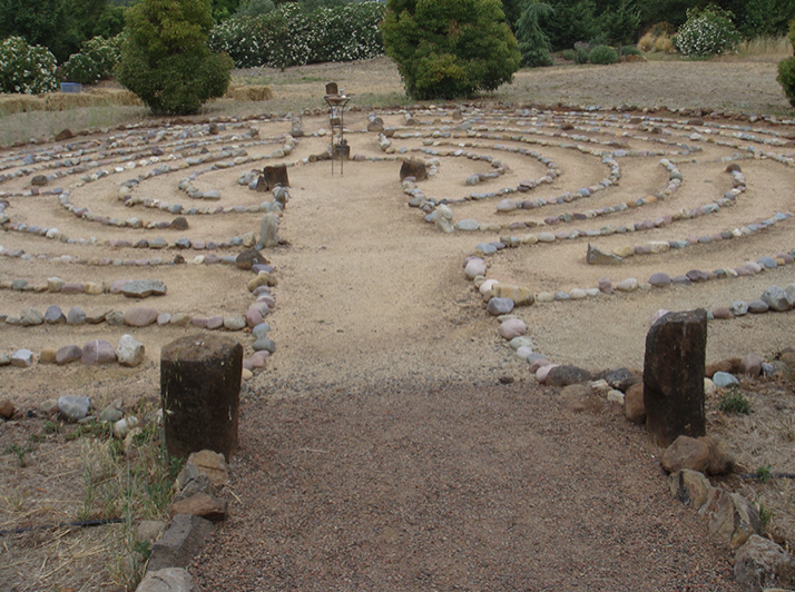 Stone labyrinth in a desert-like garden with bushes and trees in the background.